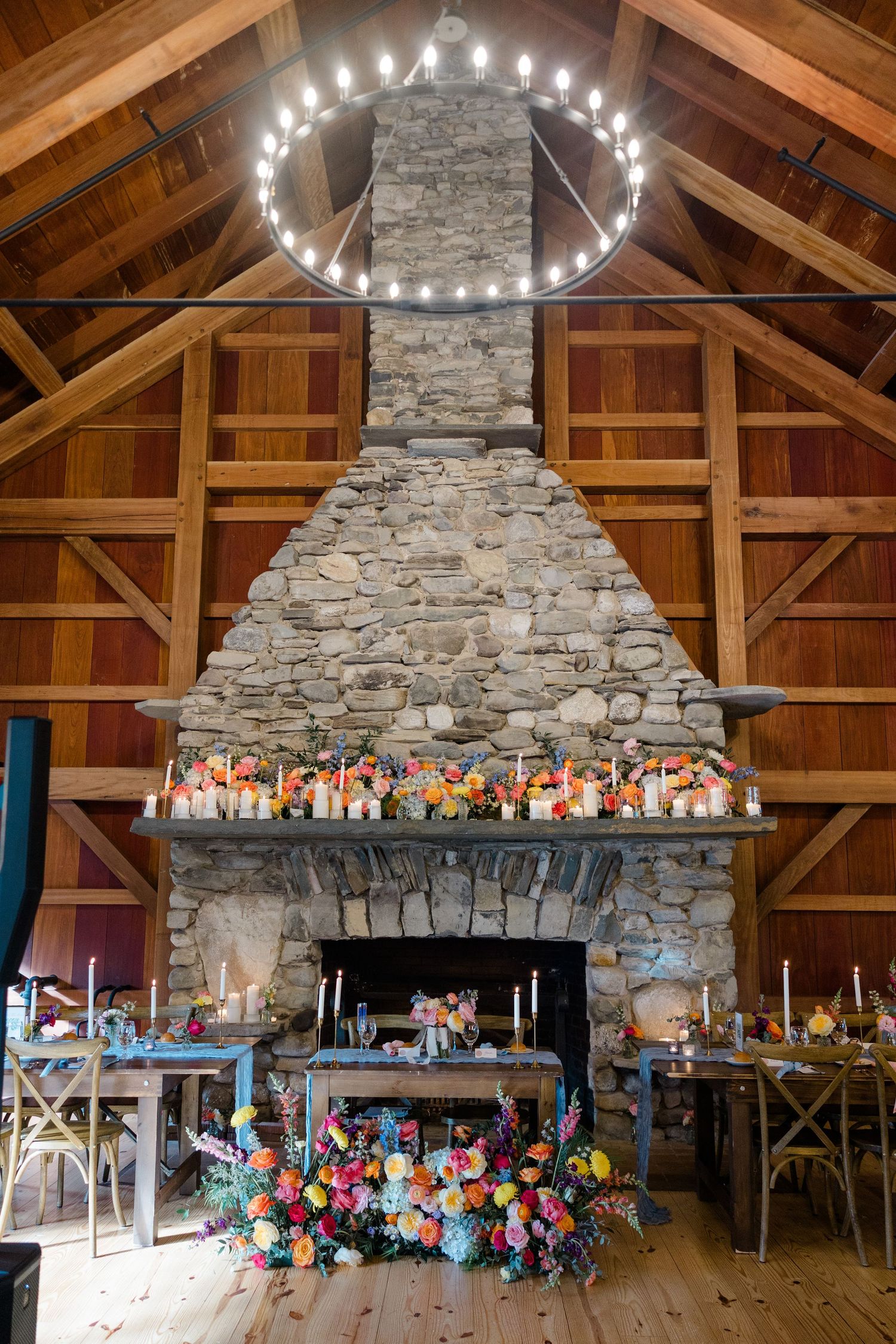 Rustic barn interior with stone fireplace, wooden beams, and circular chandelier decorated with colorful flowers.