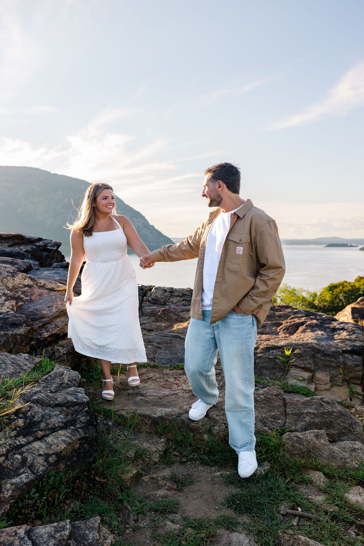 Couple holding hands on rocky overlook, woman in white dress, man in tan jacket, with mountain lake views.