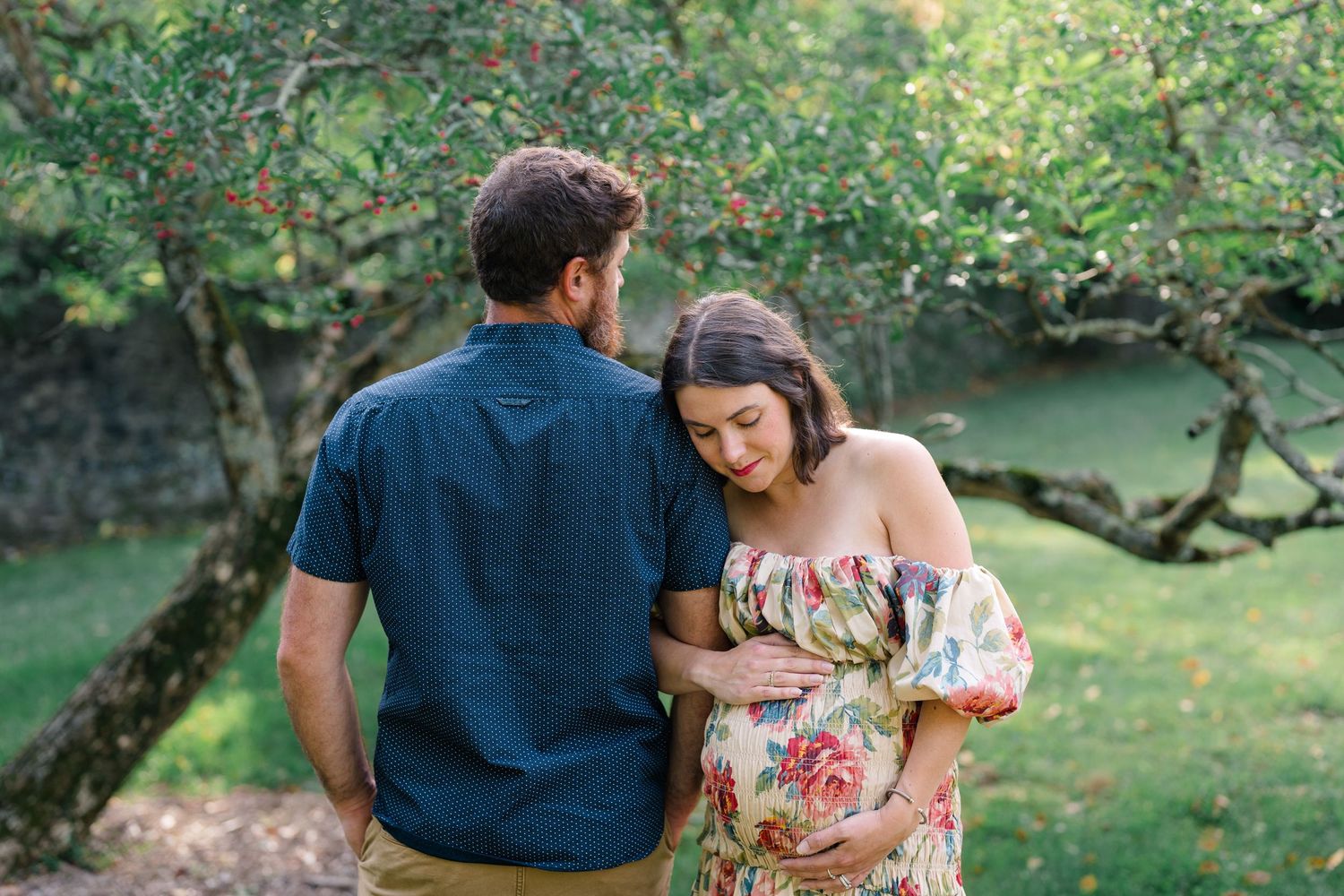 Pregnant couple embracing outdoors under apple tree, woman in floral off-shoulder dress cradling baby bump in lush green garden.