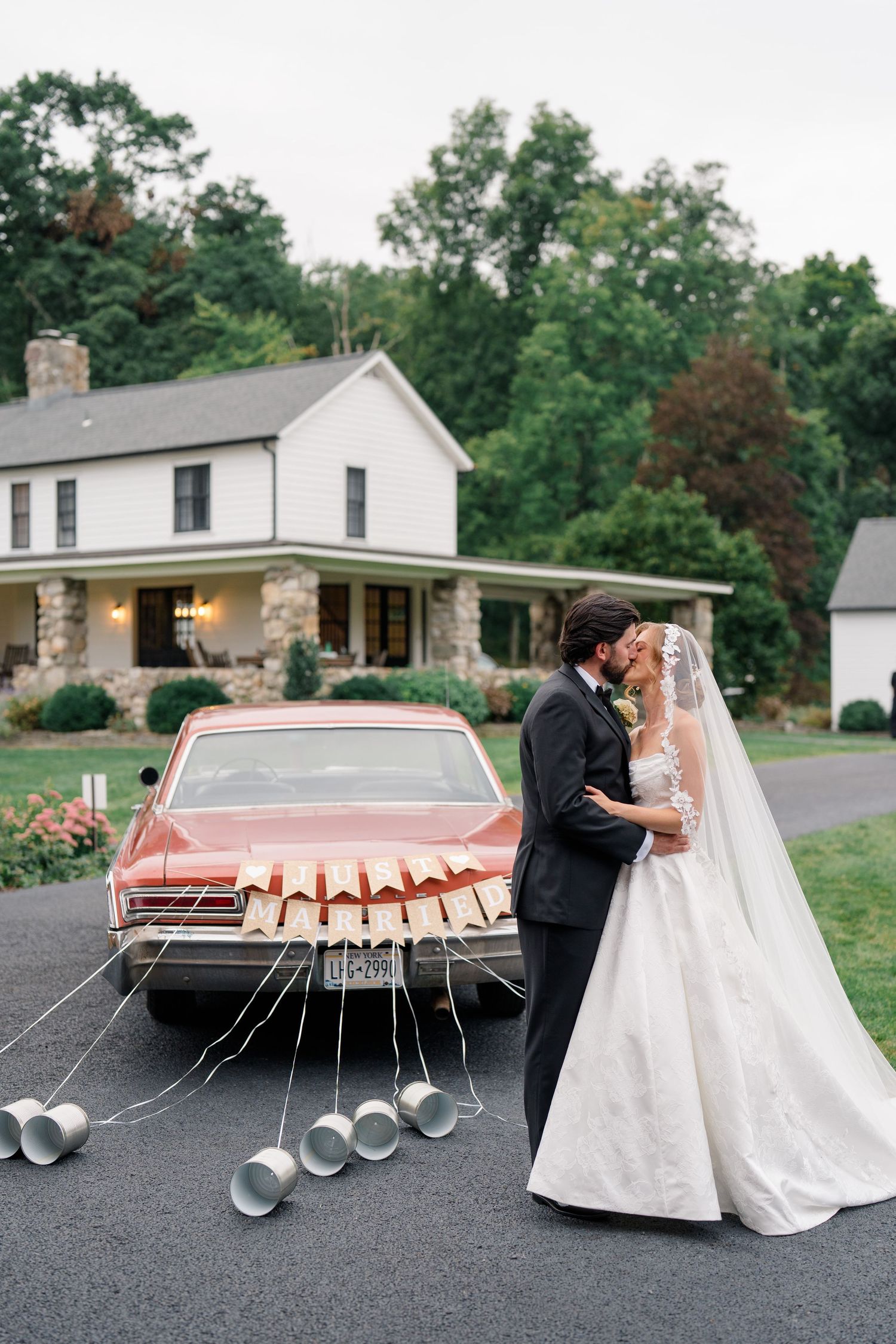 Bride and groom kissing beside a vintage red car with tin cans tied to it outside a white farmhouse.