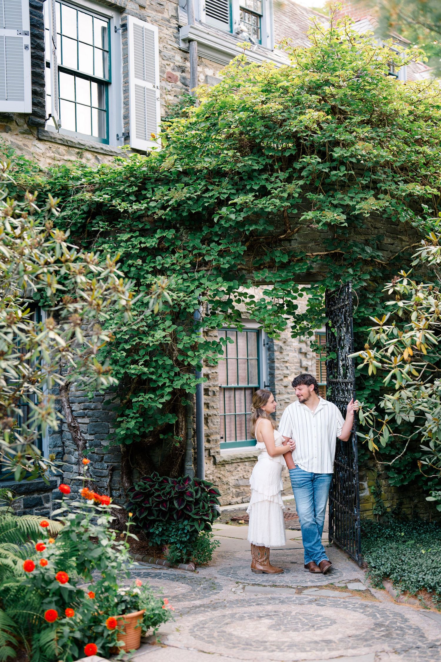 Couple embracing at ivy-covered stone building entrance with wrought iron gate and colorful garden flowers.