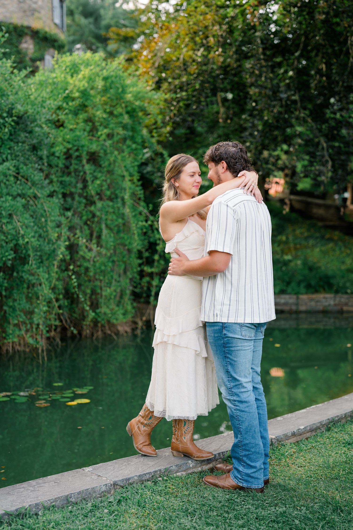 A couple embraces romantically by a serene green pond, surrounded by lush garden greenery during an outdoor photo session.