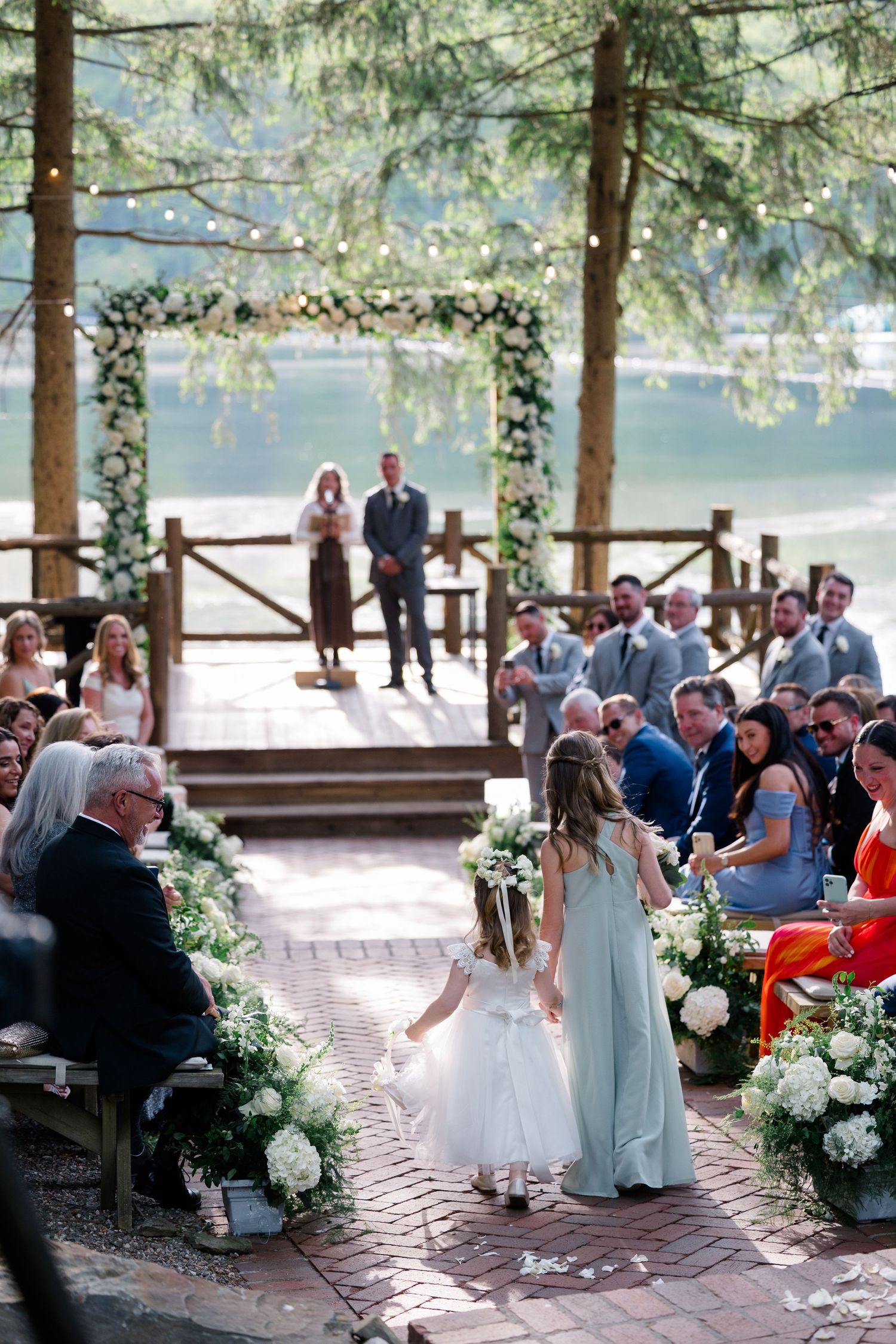 Outdoor wedding ceremony under pine trees with floral arch and brick pathway.