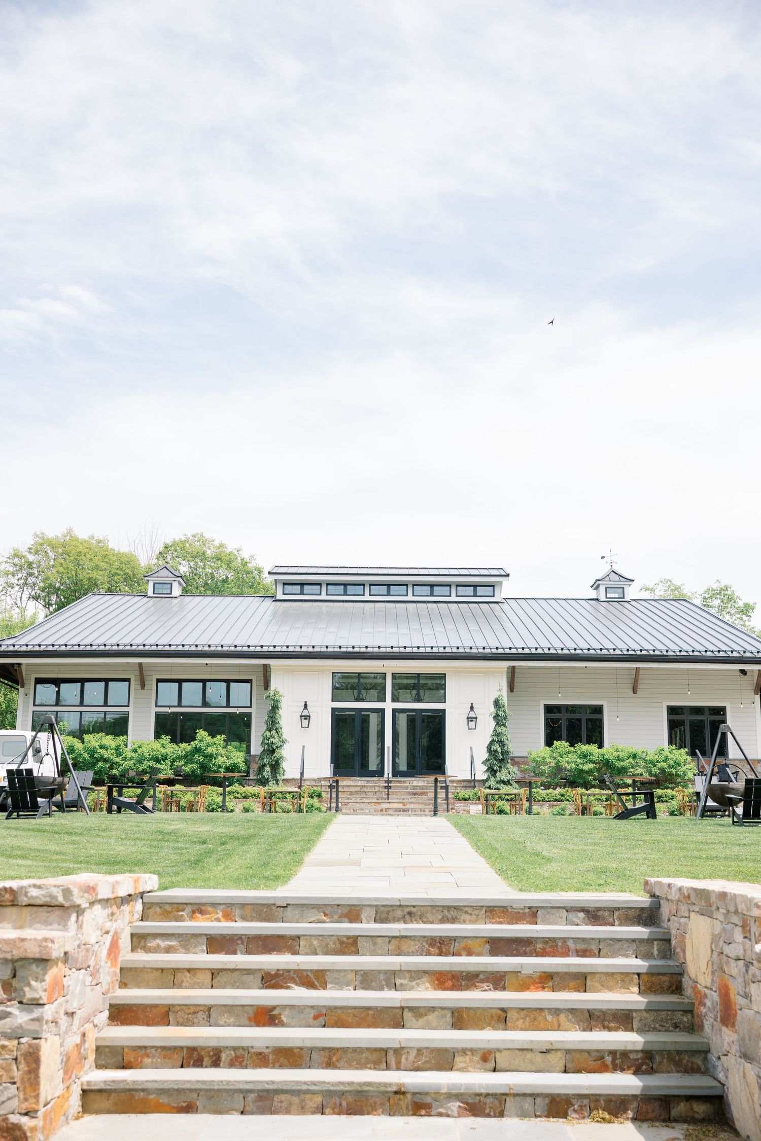 Modern event venue building with stone steps and green landscaping features a metal roof and symmetrical design.