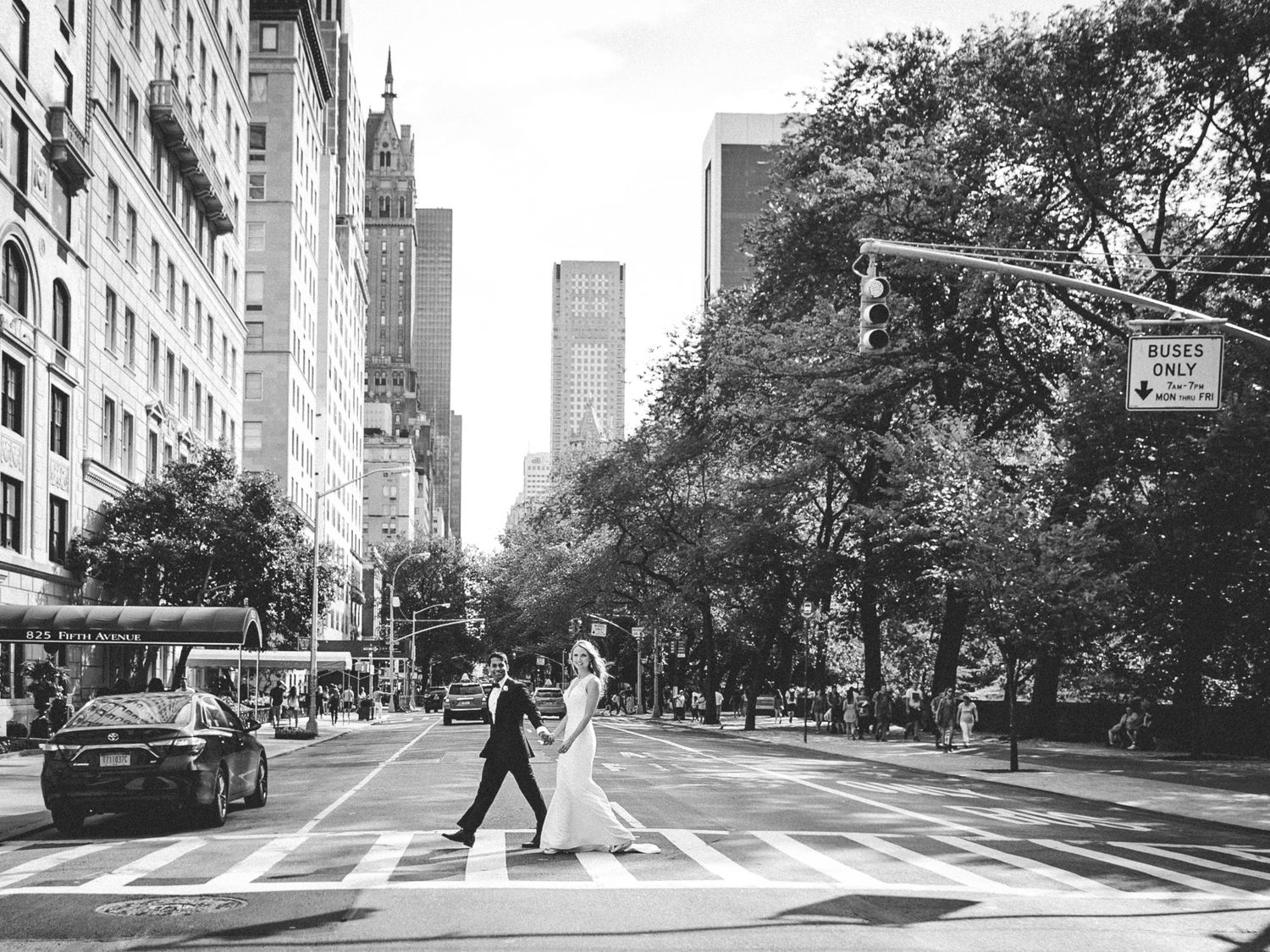 A black and white cityscape of New York City with pedestrians crossing a crosswalk on a tree-lined urban street.