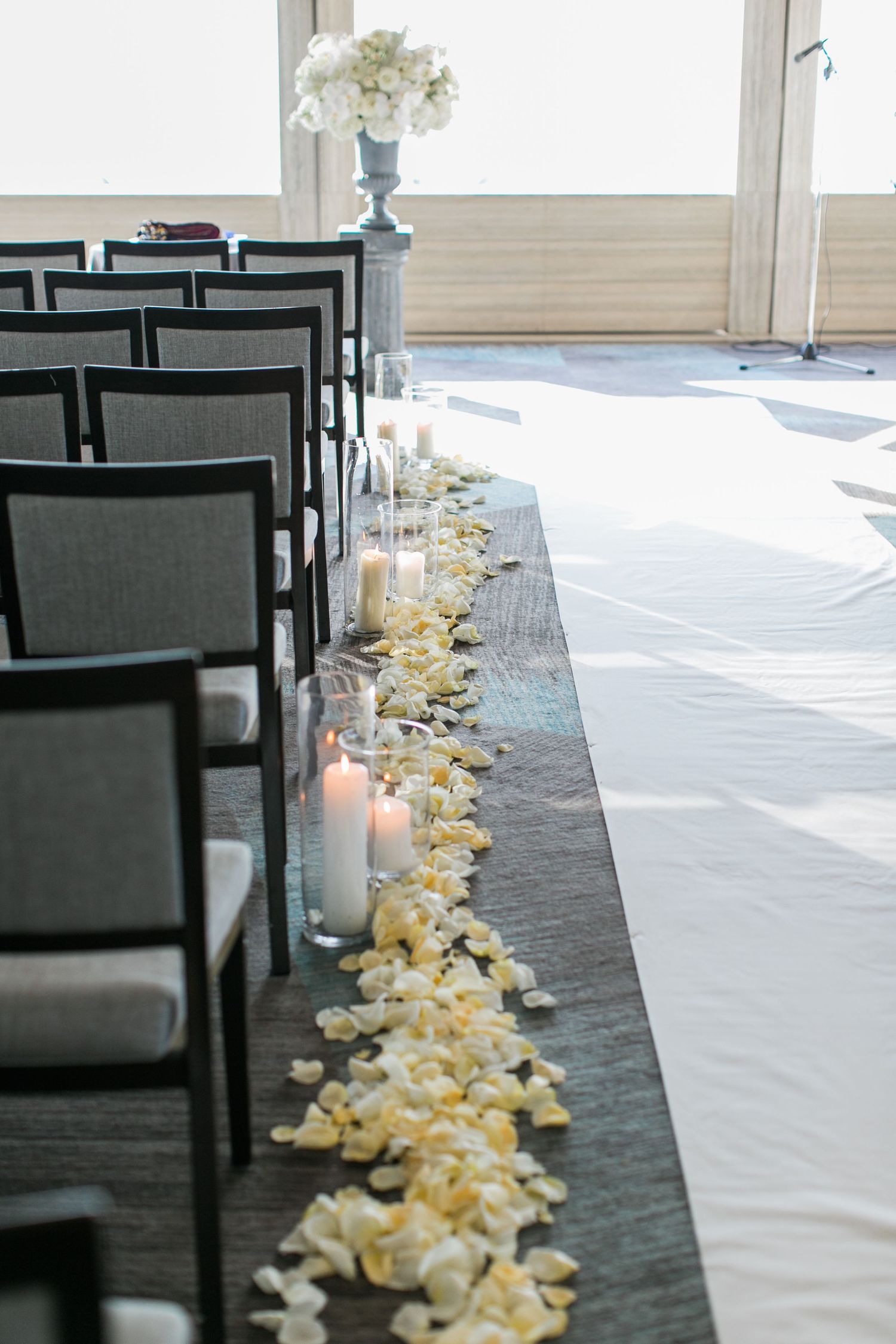 A wedding ceremony aisle decorated with white flower petals and candles alongside black chairs.