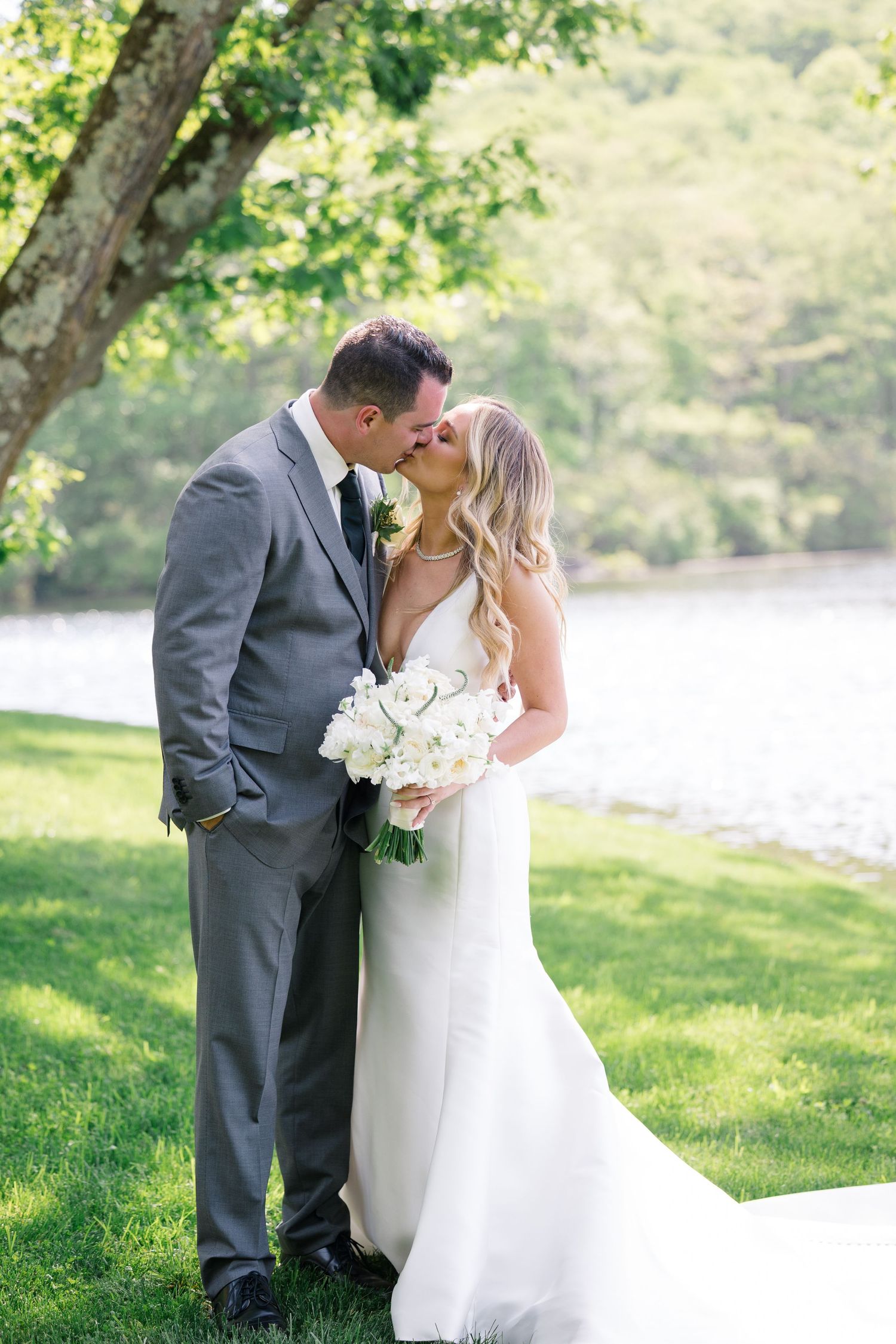 A romantic wedding couple shares a kiss by a lake on a sunny day, with the bride holding a white floral bouquet.