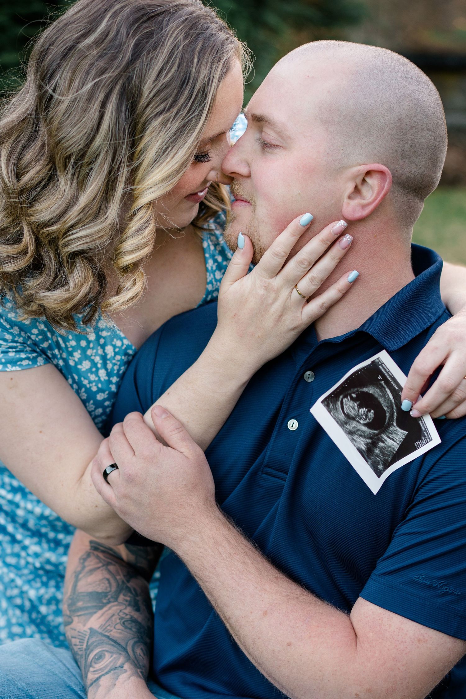 An intimate couple's portrait shows them embracing while holding an ultrasound photo in an outdoor setting.
