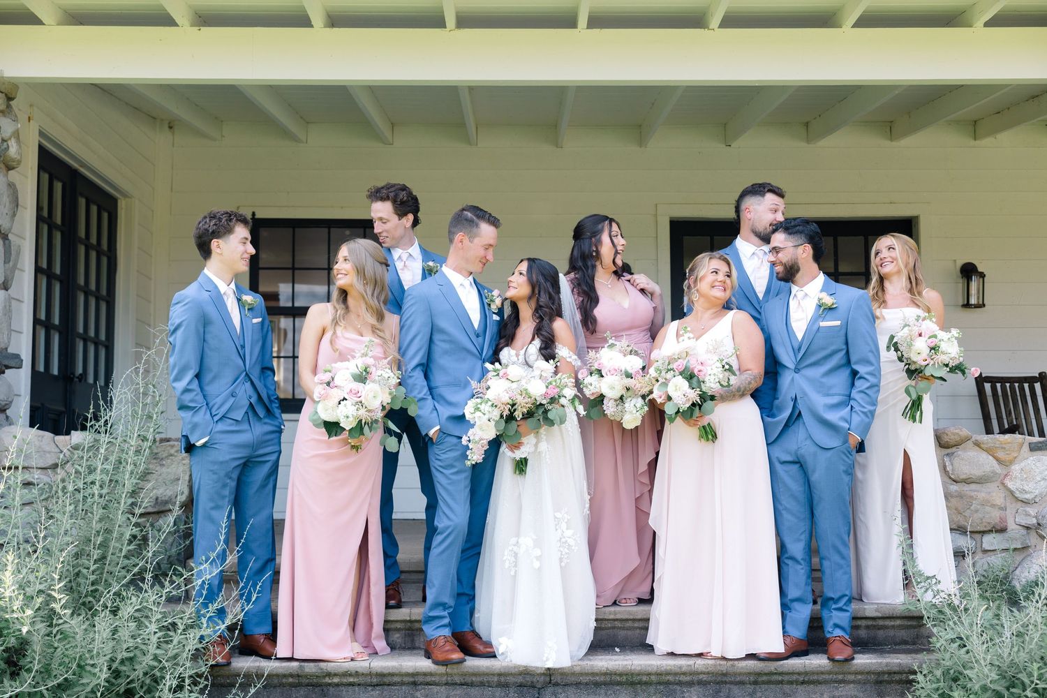Wedding party in coordinating blue suits and pink dresses poses on covered porch.