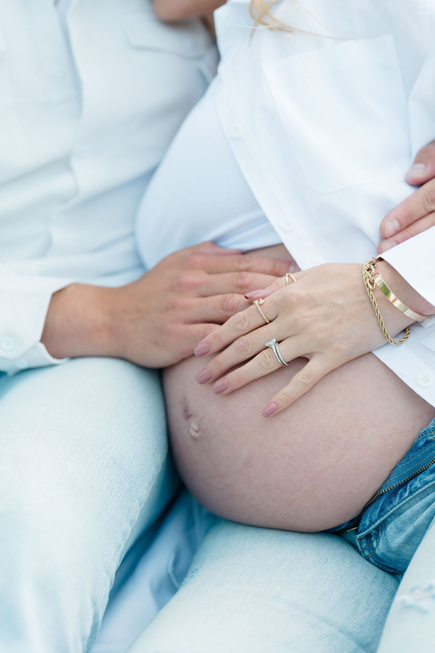 Close up shots of hands resting on a pregnant belly in a white top show delicate rings and tender moments.