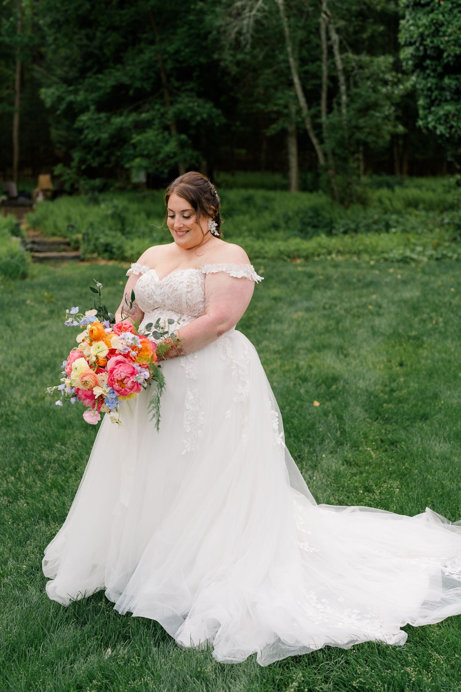A bride in a flowing white gown poses gracefully on grassy lawn surrounded by greenery.