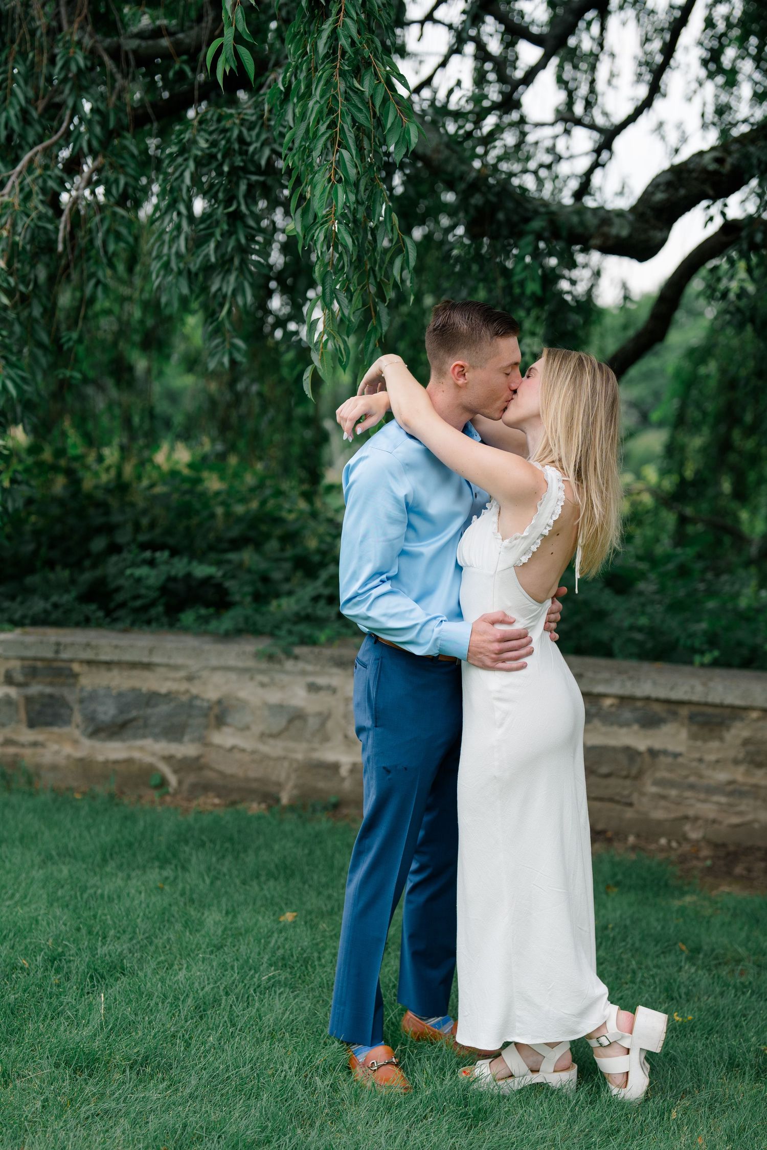 Couple sharing a romantic kiss under a weeping willow tree, groom in blue suit, bride in white dress.