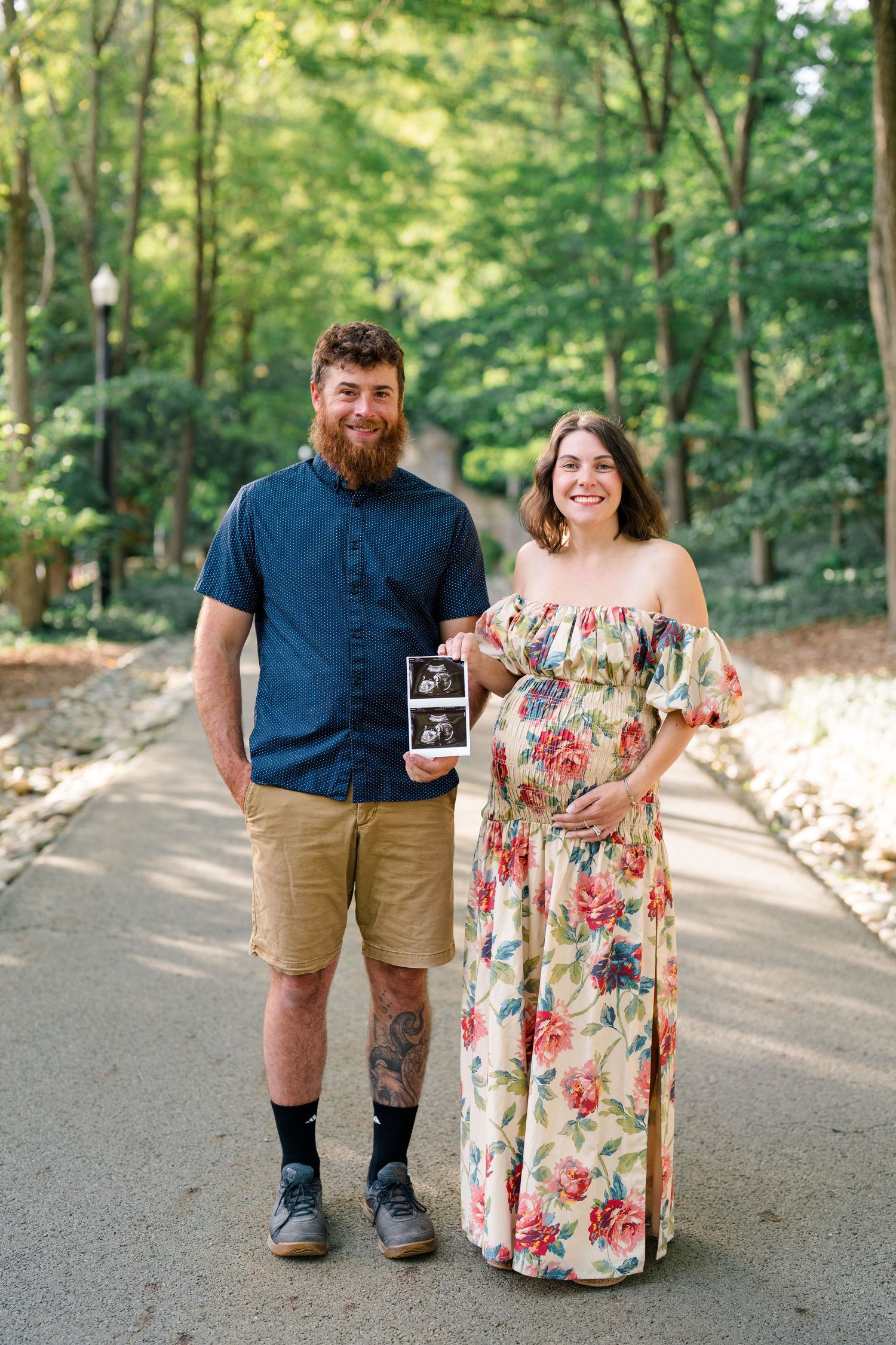 Pregnant couple holding ultrasound photo outdoors on a tree-lined path, woman in floral dress cradling baby bump.