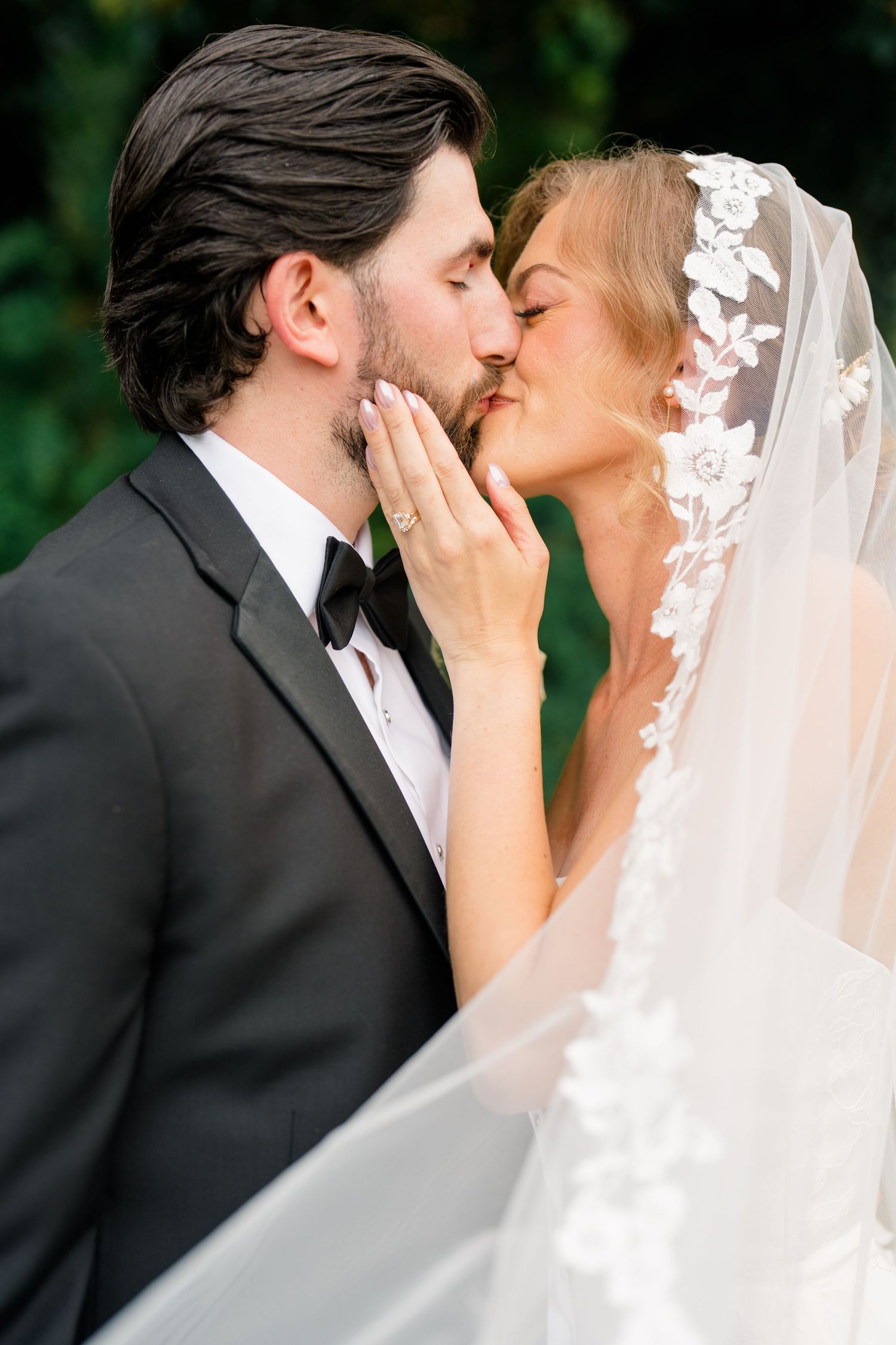 Bride and groom share a tender kiss, bride's hand on groom's face, lace floral veil draped elegantly.