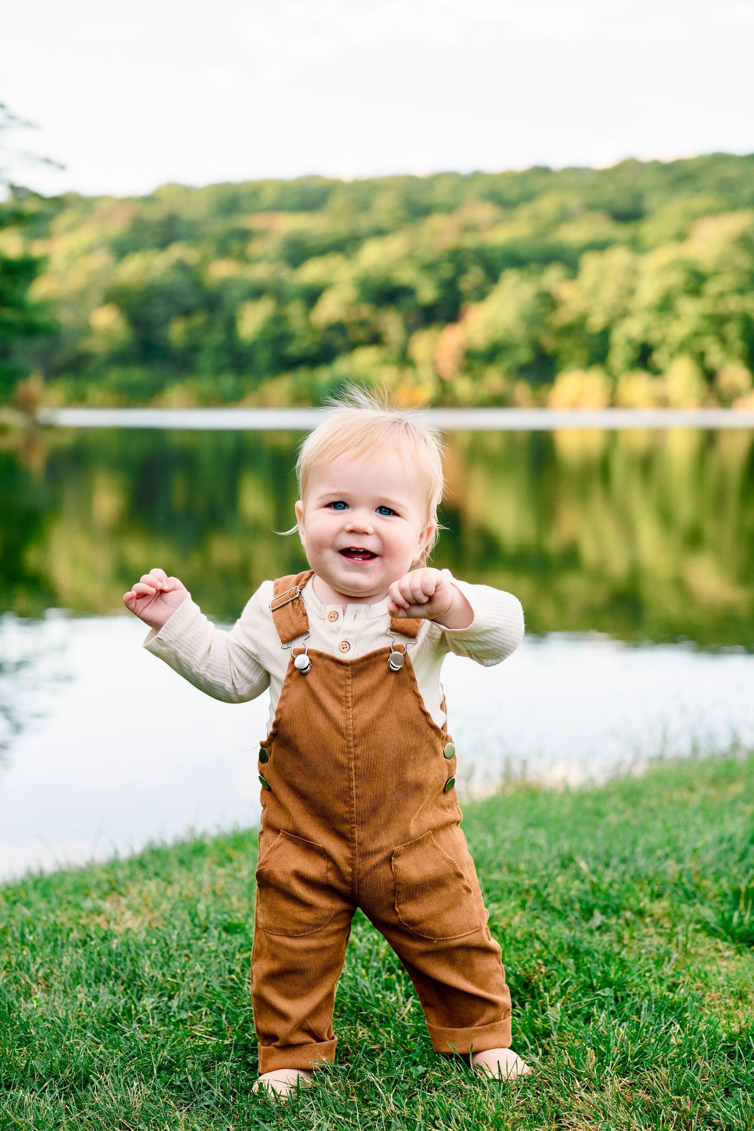 A happy baby in brown overalls and white shirt stands on green grass near a reflective lake surrounded by trees.
