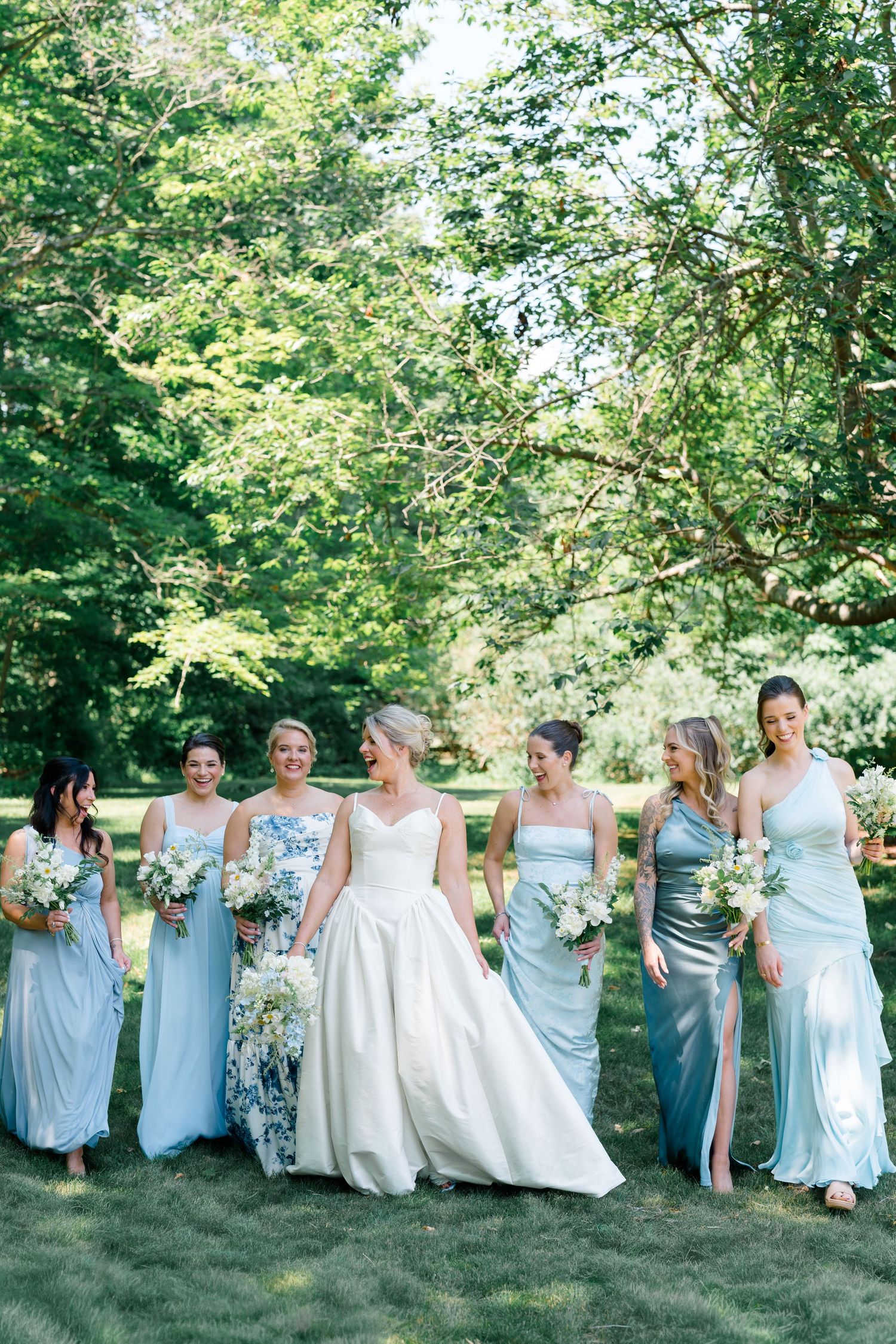Bride in white ball gown walks with six bridesmaids in mismatched blue and silver dresses in lush garden.