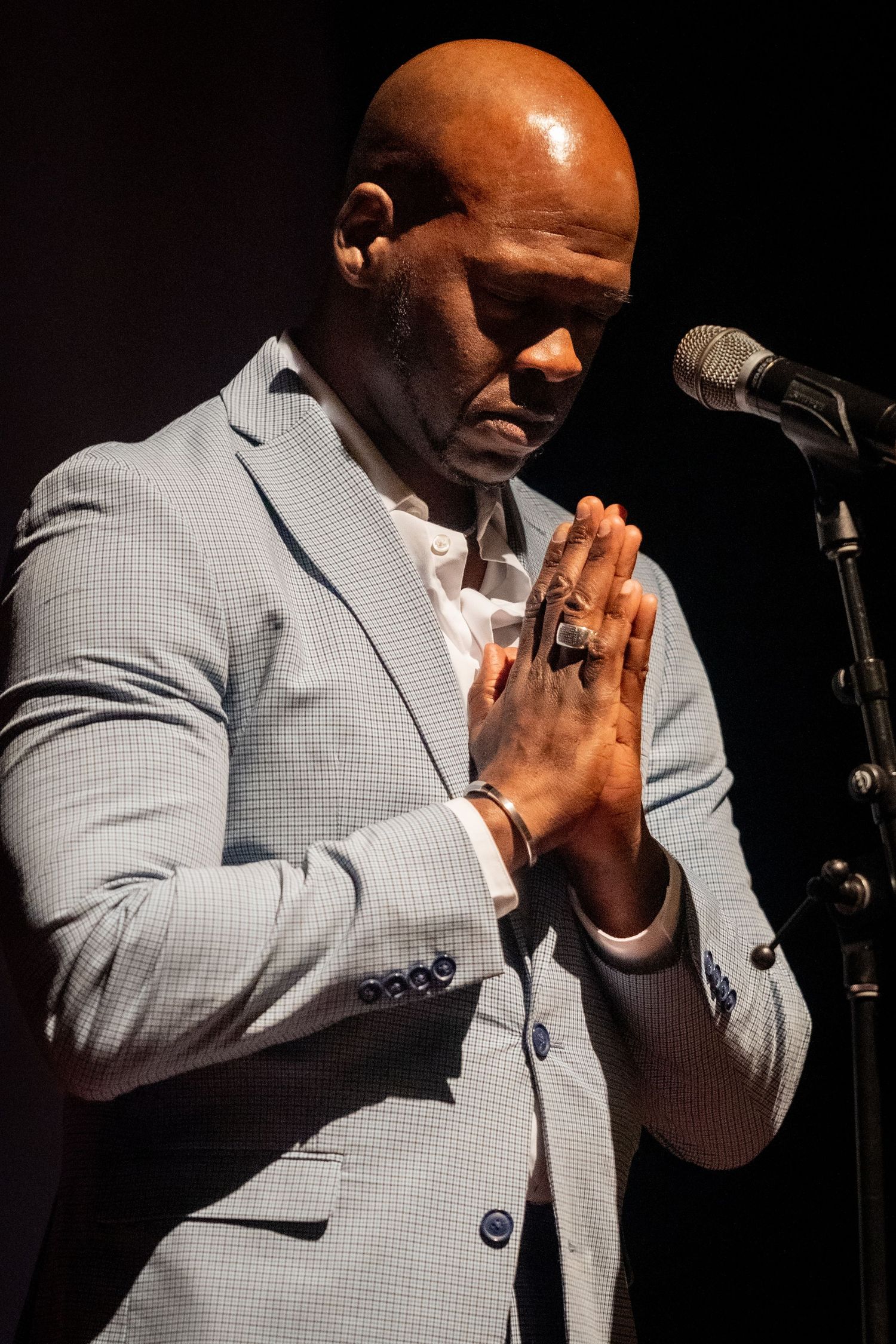 Performer in light gray suit jacket clasps hands in prayer position on dark stage.