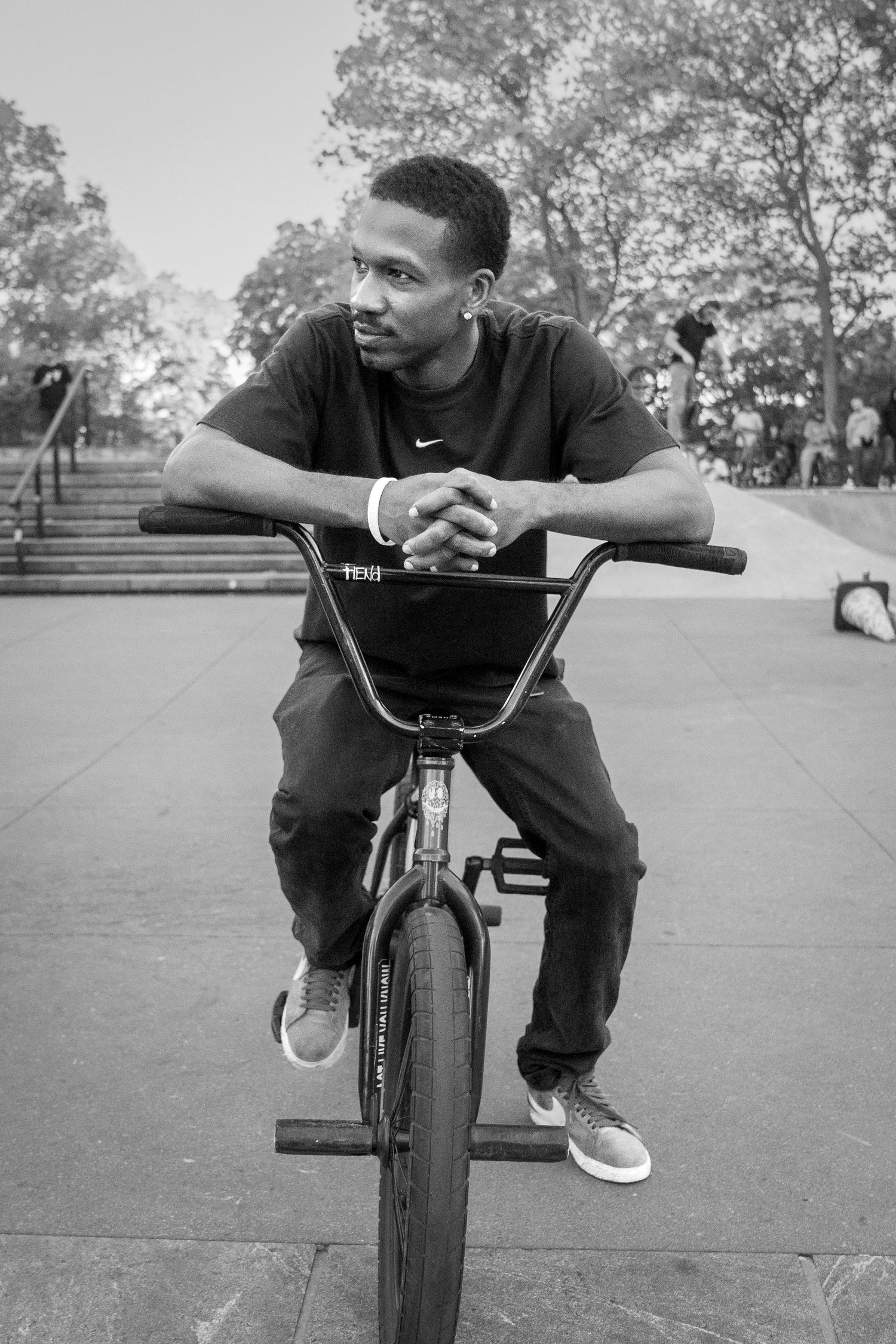 BMX rider in casual wear resting on handlebars in an urban park setting.