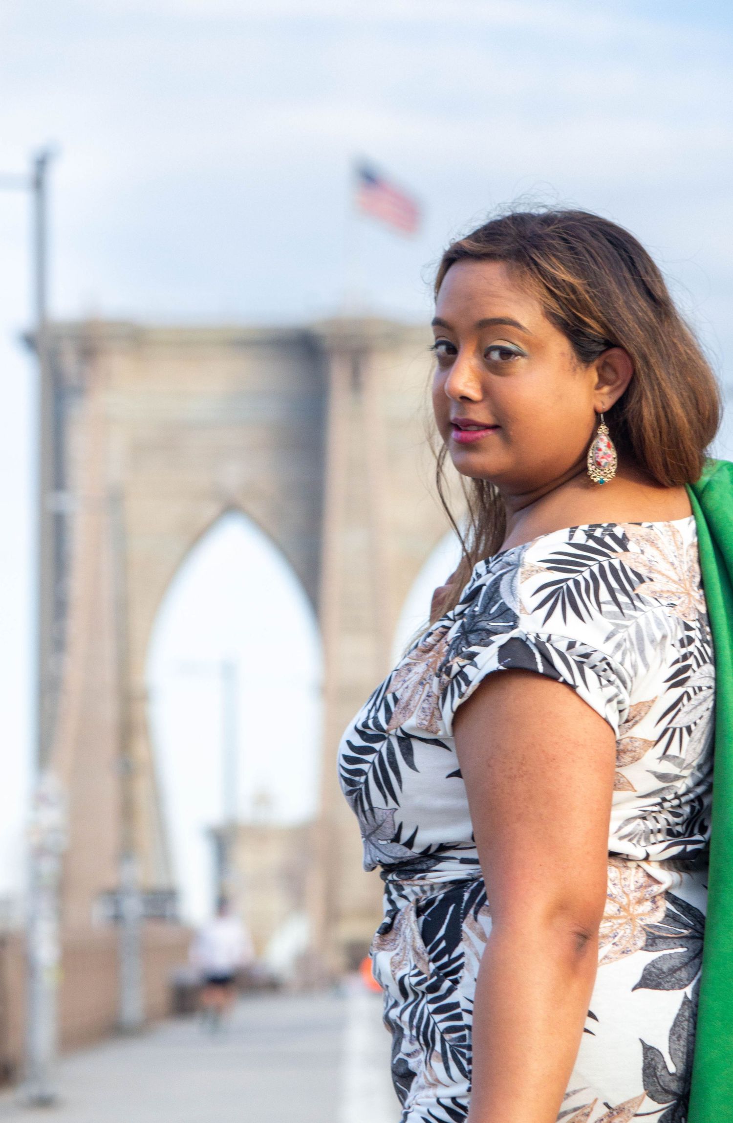 Woman in floral dress poses on Brooklyn Bridge with American flag in background.