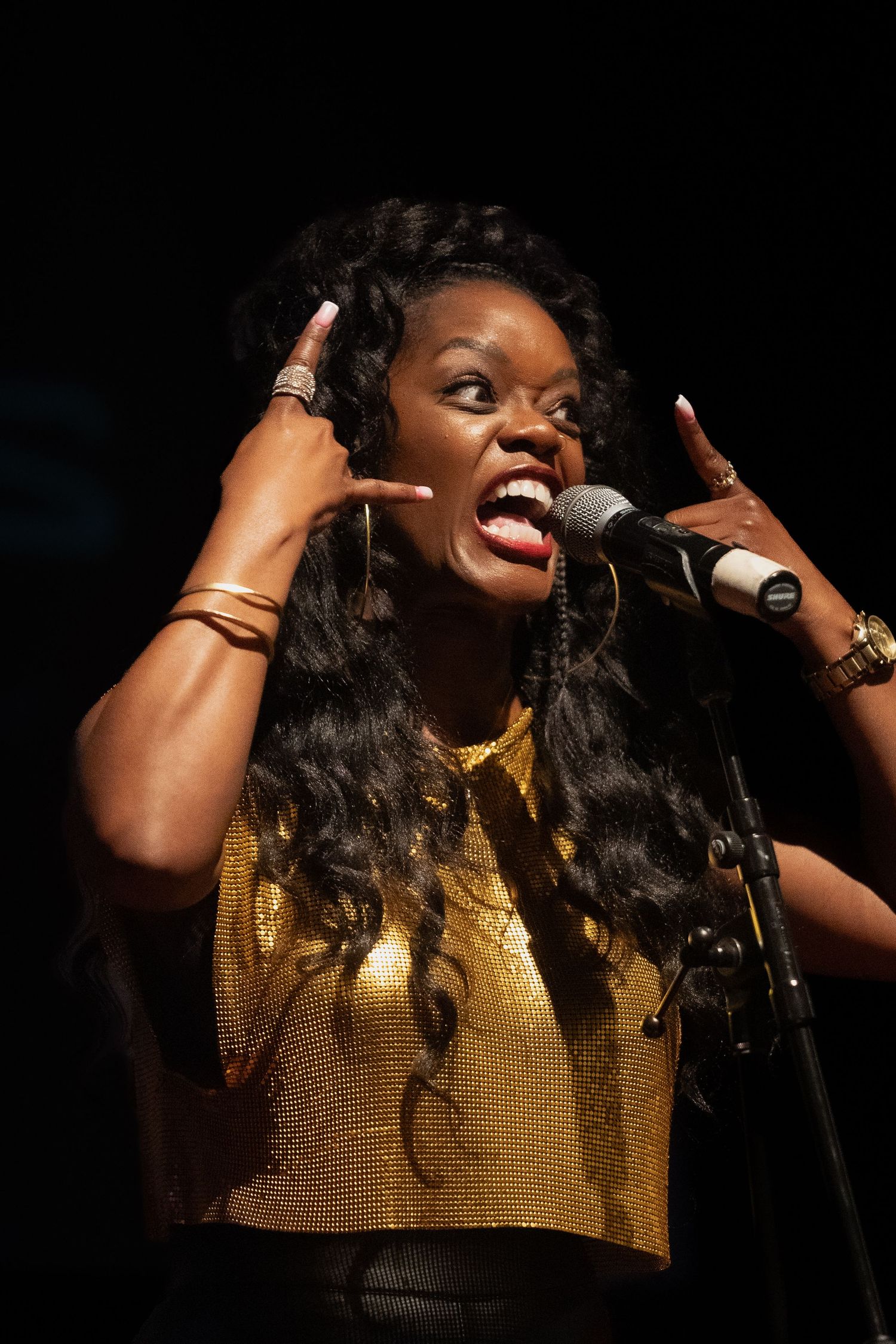 Singer in gold top performs energetically with microphone on dark stage.