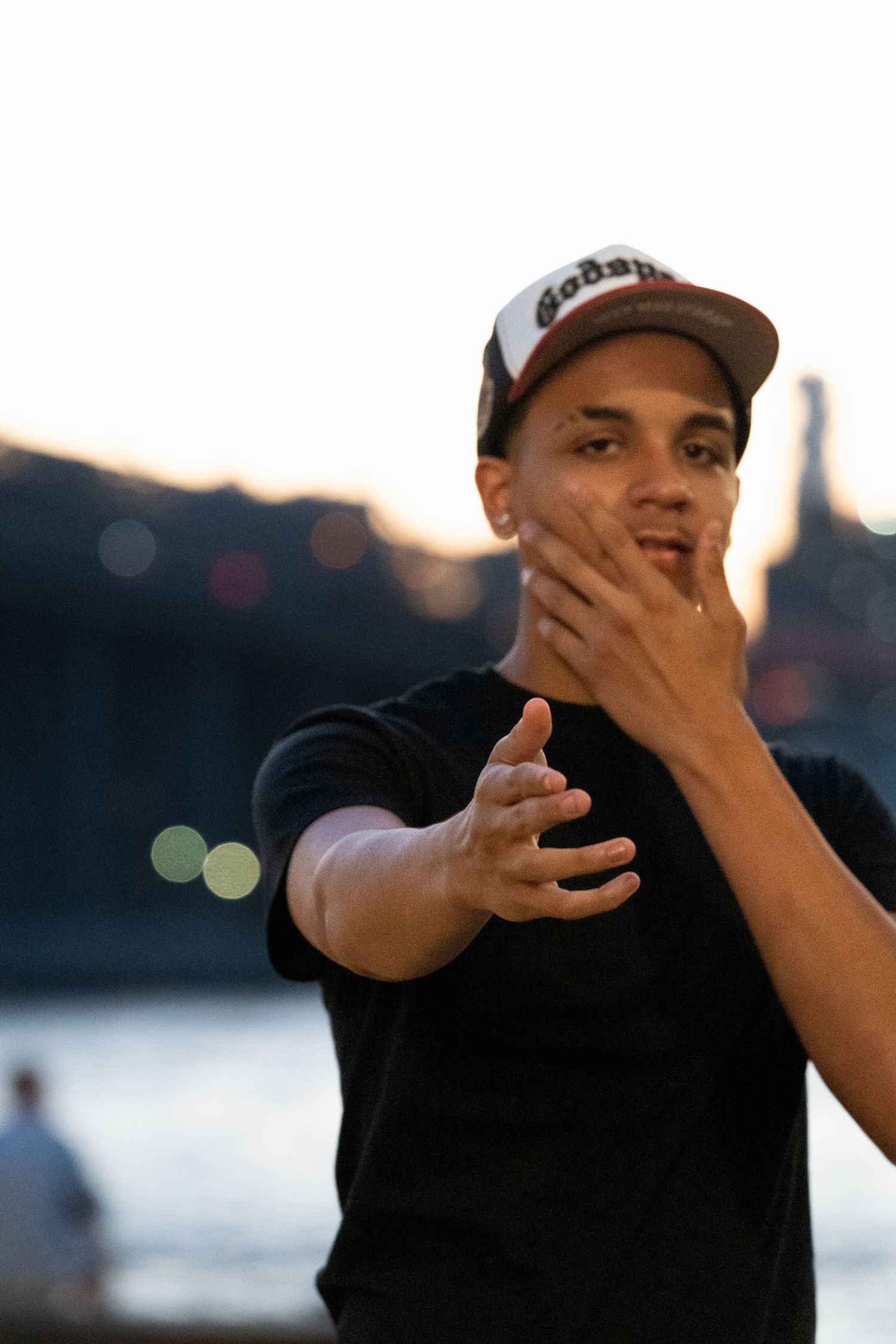 A person in a baseball cap gestures expressively against a sunset waterfront backdrop.