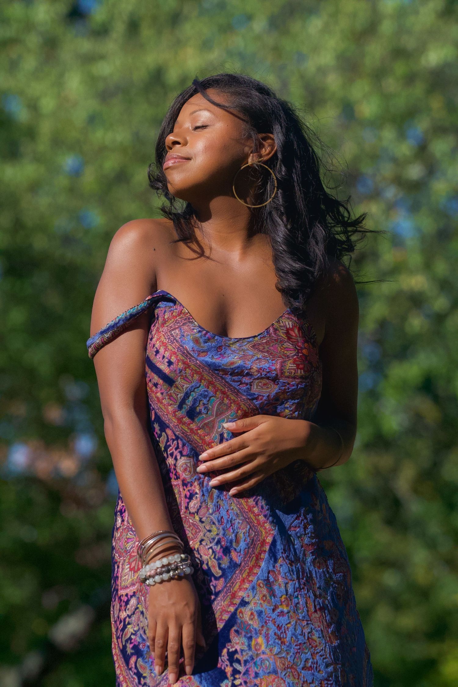 Person in patterned purple dress poses gracefully outdoors with natural greenery.