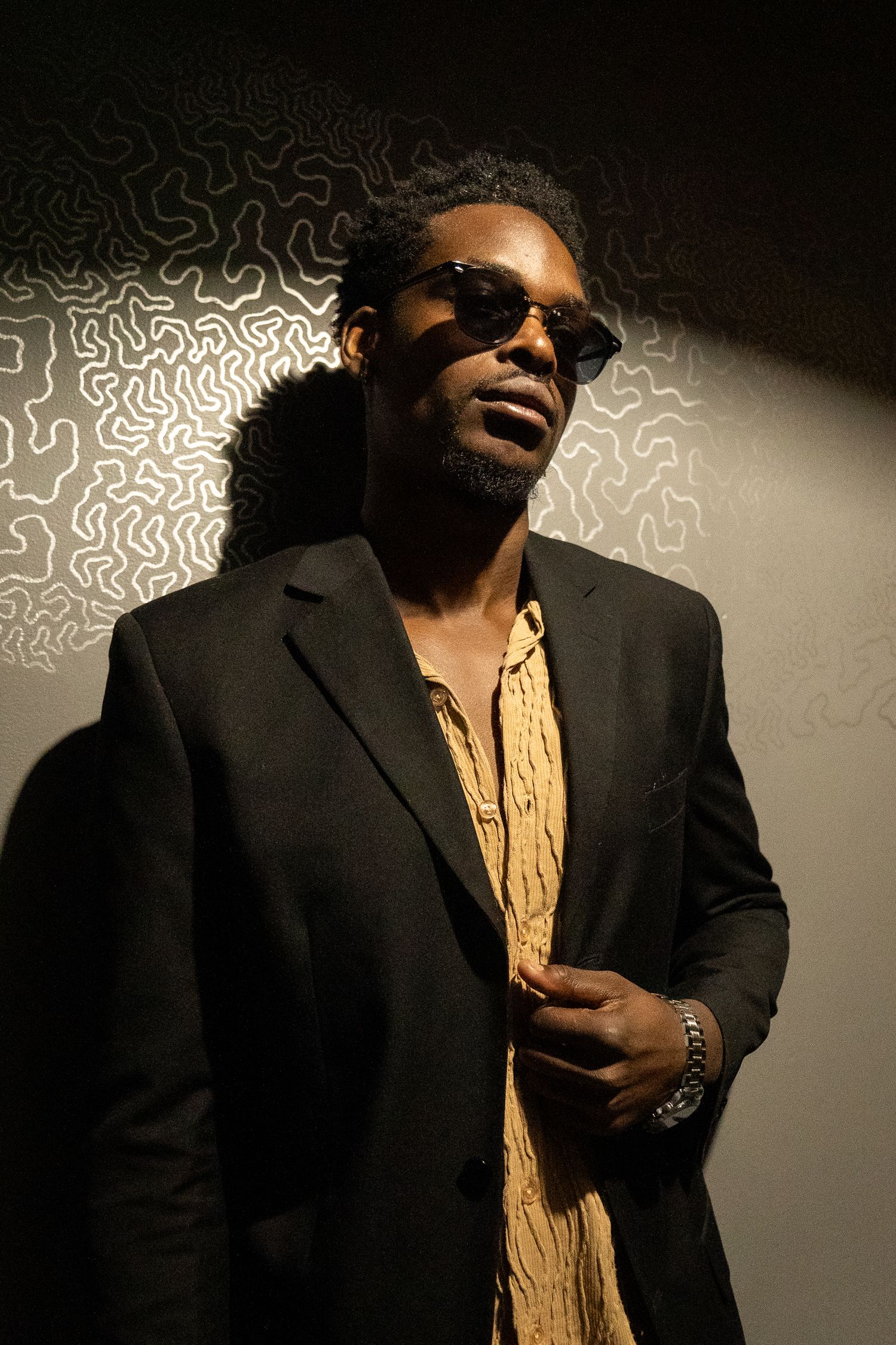A stylish performer in a black suit and gold scarf strikes dramatic poses against a light background in a photo series.