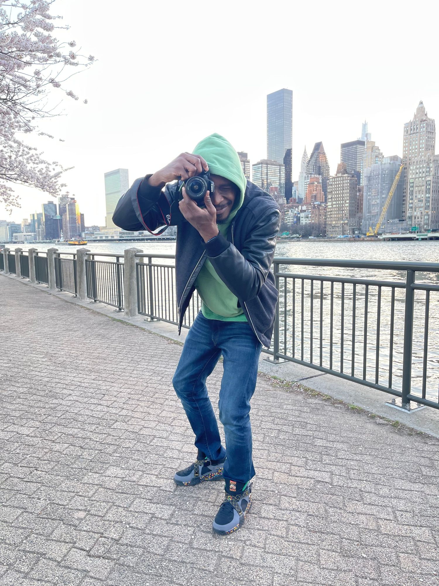 Person in casual wear dancing on a waterfront promenade with NYC skyline in background.