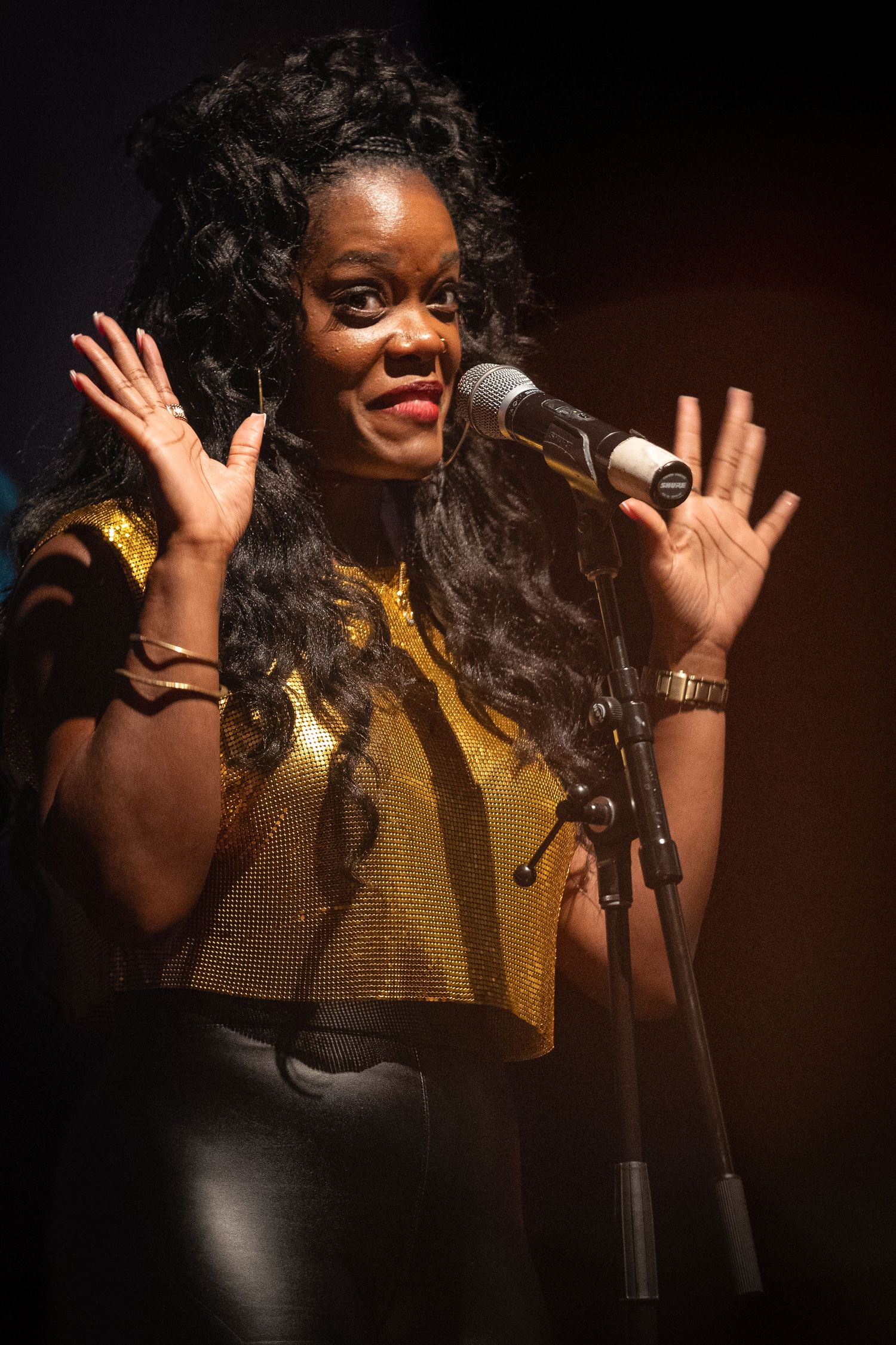 Performer in golden top singing energetically into microphone on darkened stage.