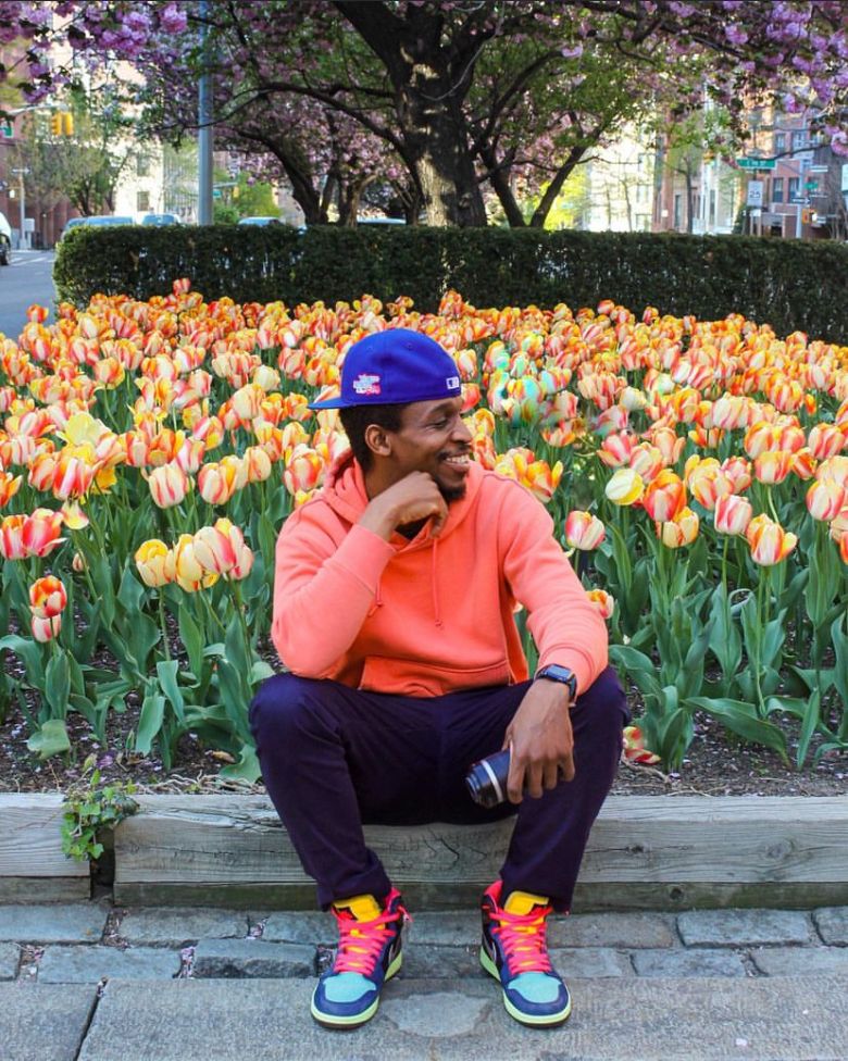 Person sitting among vibrant orange and yellow tulips wearing bright coral hoodie and blue cap.