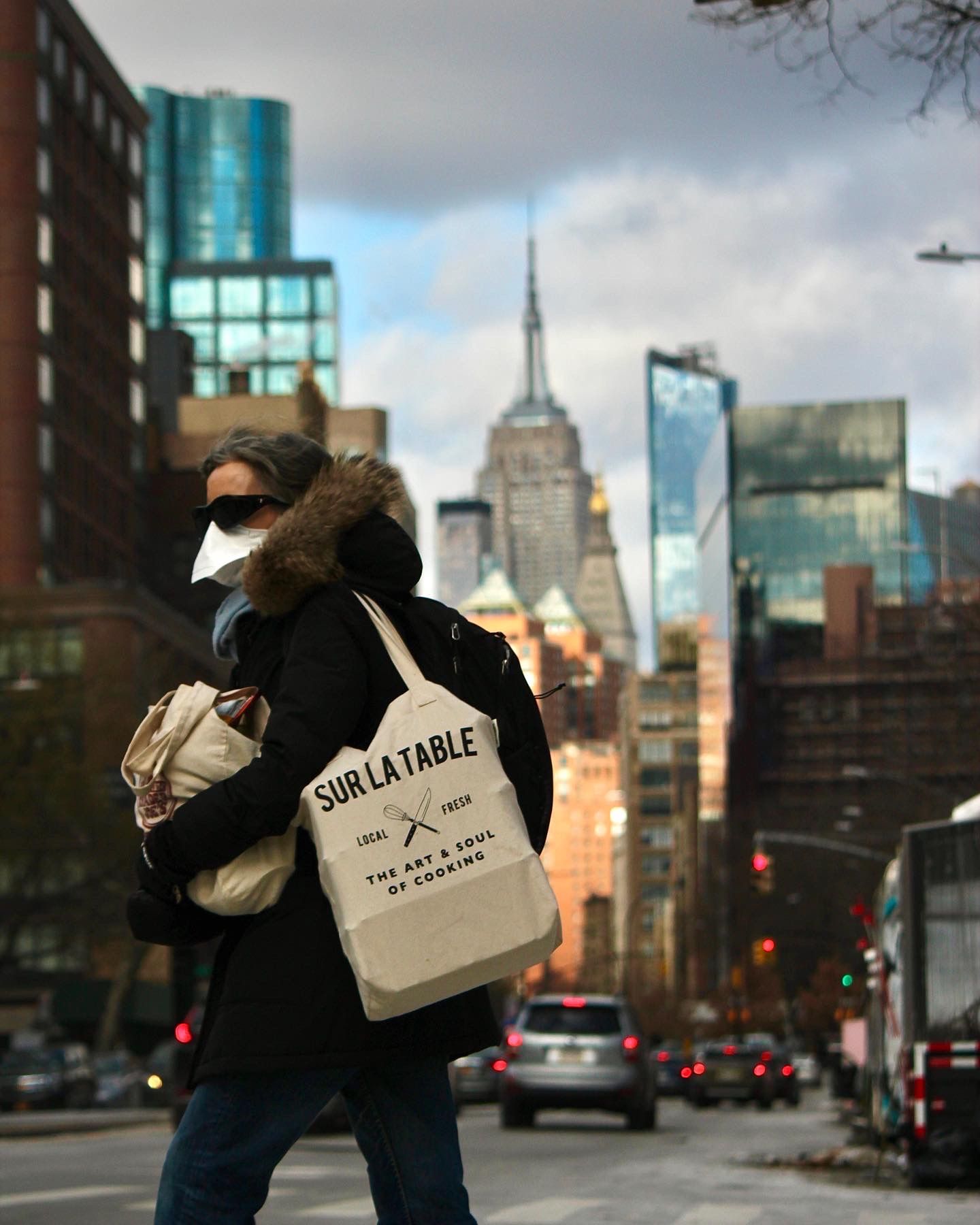 Person wearing winter coat and mask carries tote bag on NYC street with Empire State Building visible.