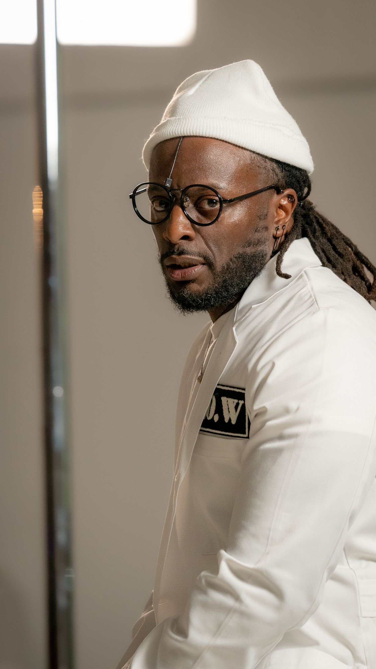 Person in white outfit and glasses poses against light background wearing beanie.
