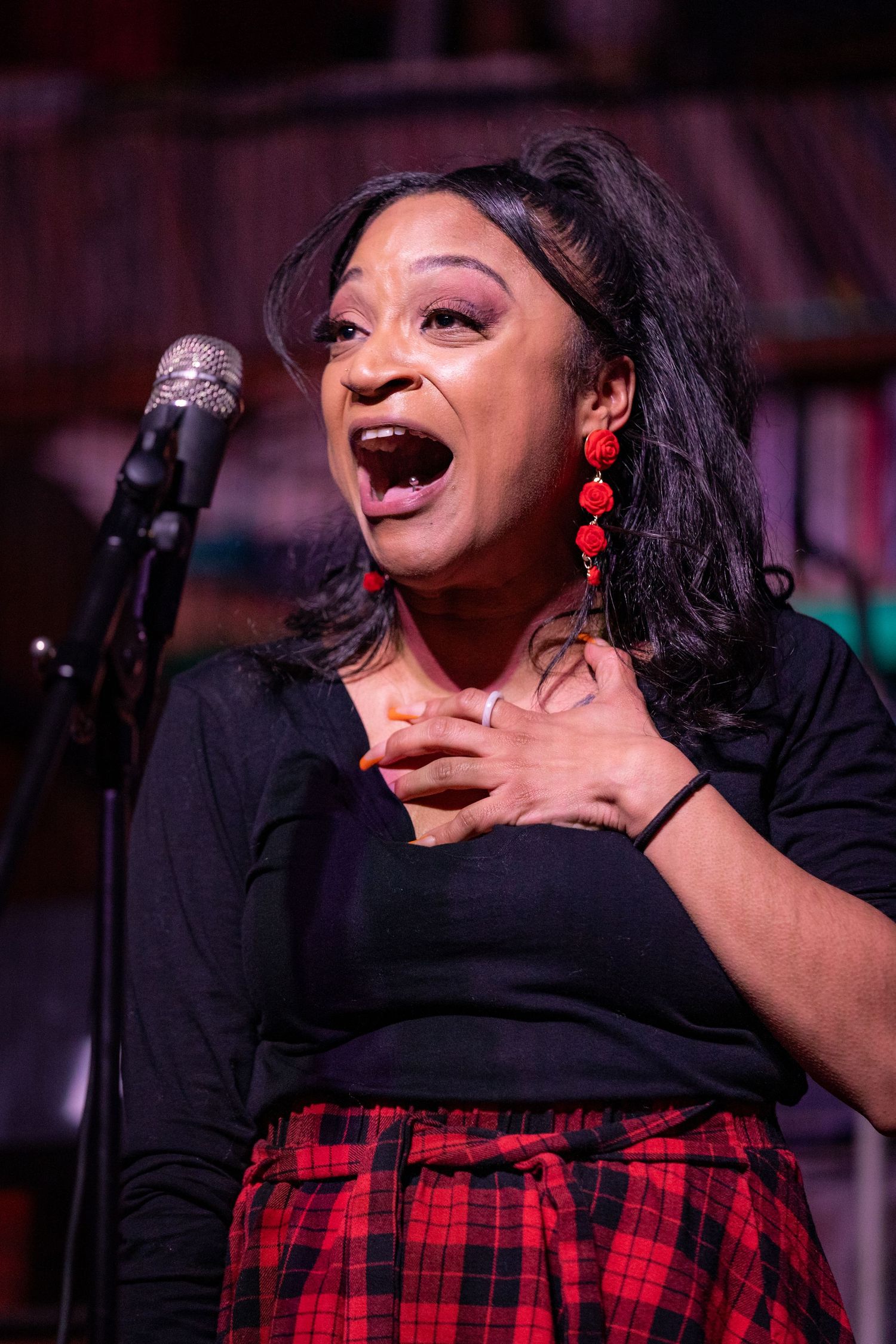 Performer in black top and plaid skirt singing emotively into microphone on dimly lit stage.