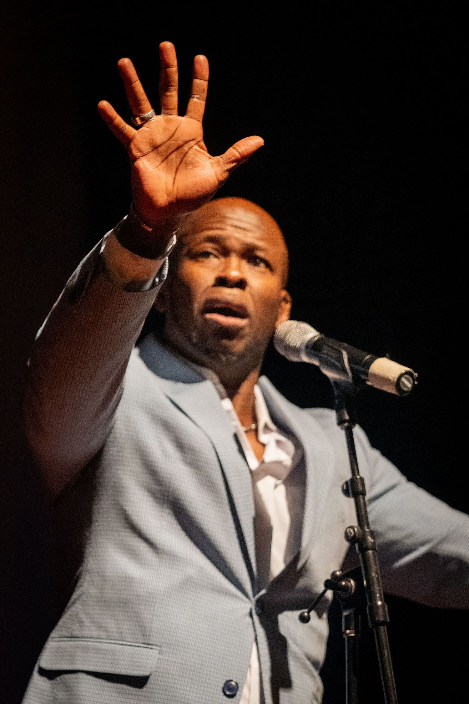 Performer in gray suit singing passionately into microphone with raised hands.