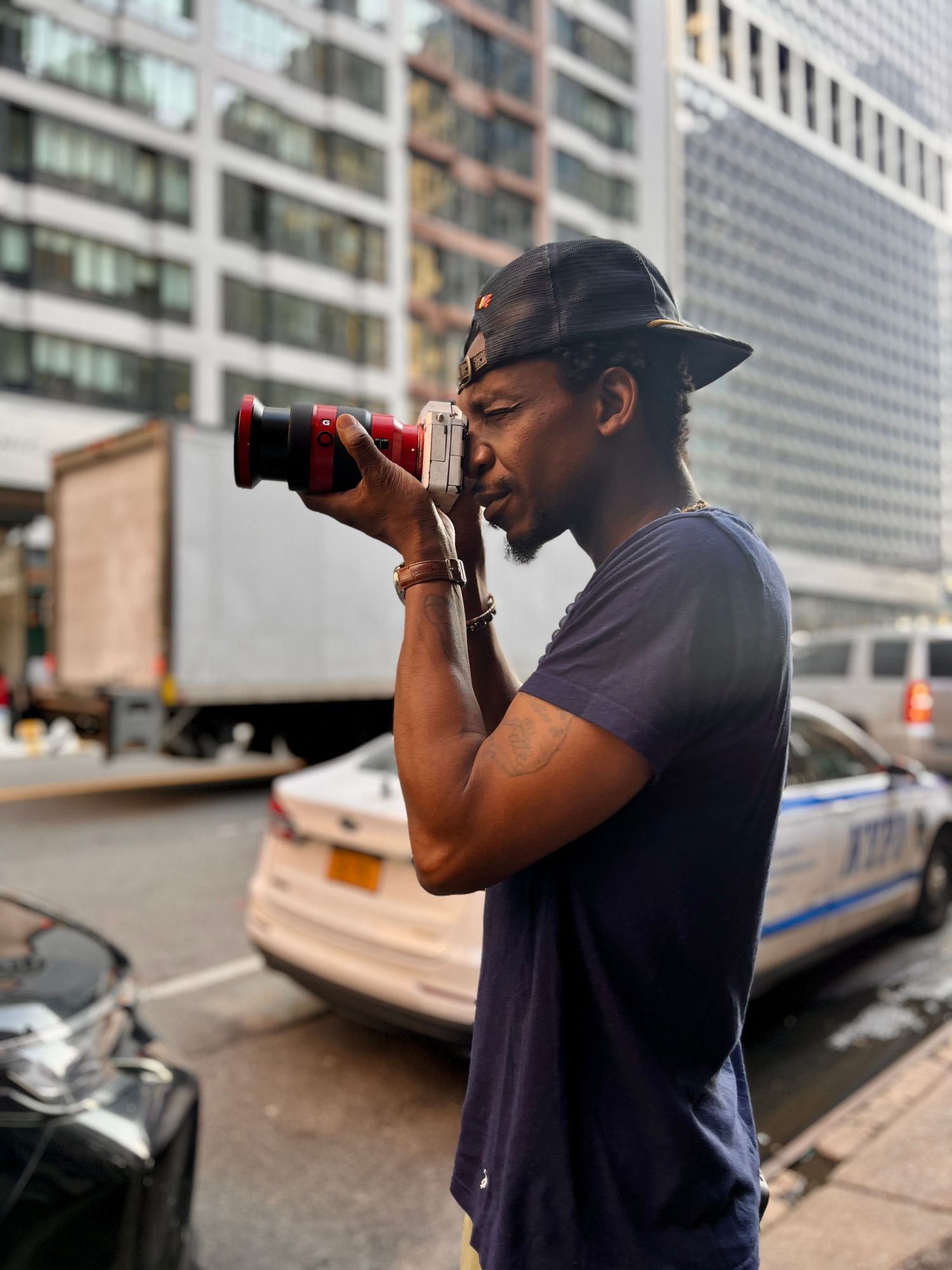 A photographer in a navy shirt takes pictures from different angles on a city street with tall buildings in the background.