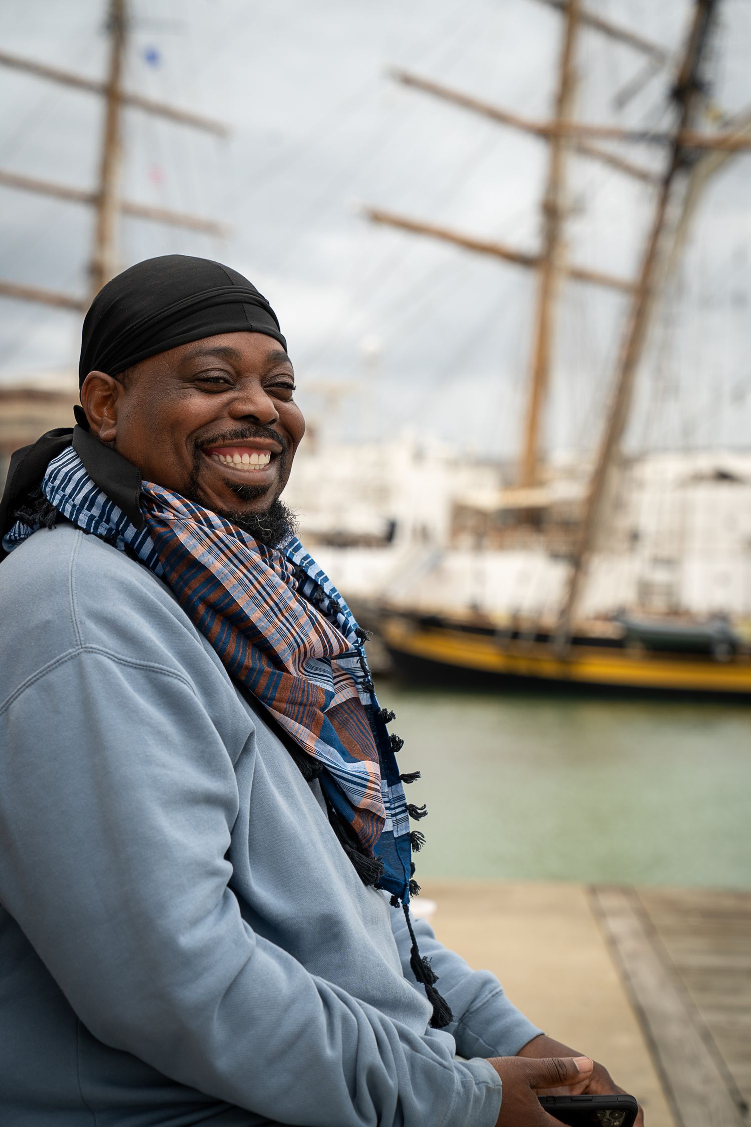 Person in grey coat and scarf smiling while sitting by a harbor with sailing ships in background.