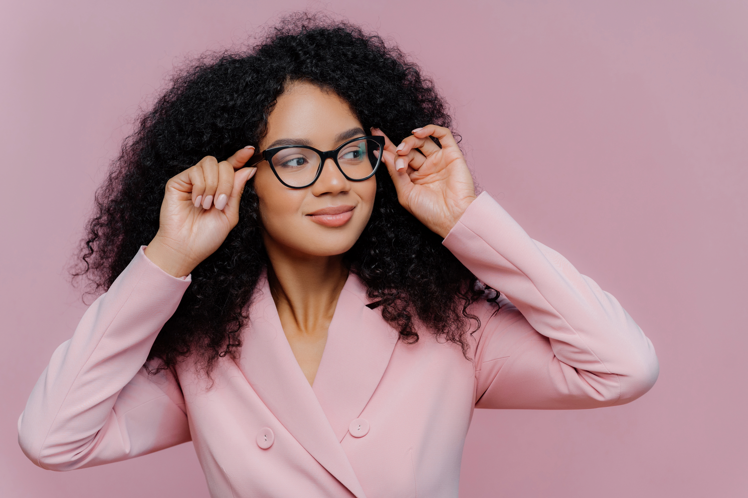 A stylish woman with curly hair wears black-framed glasses and a pink blazer while adjusting her frames.
