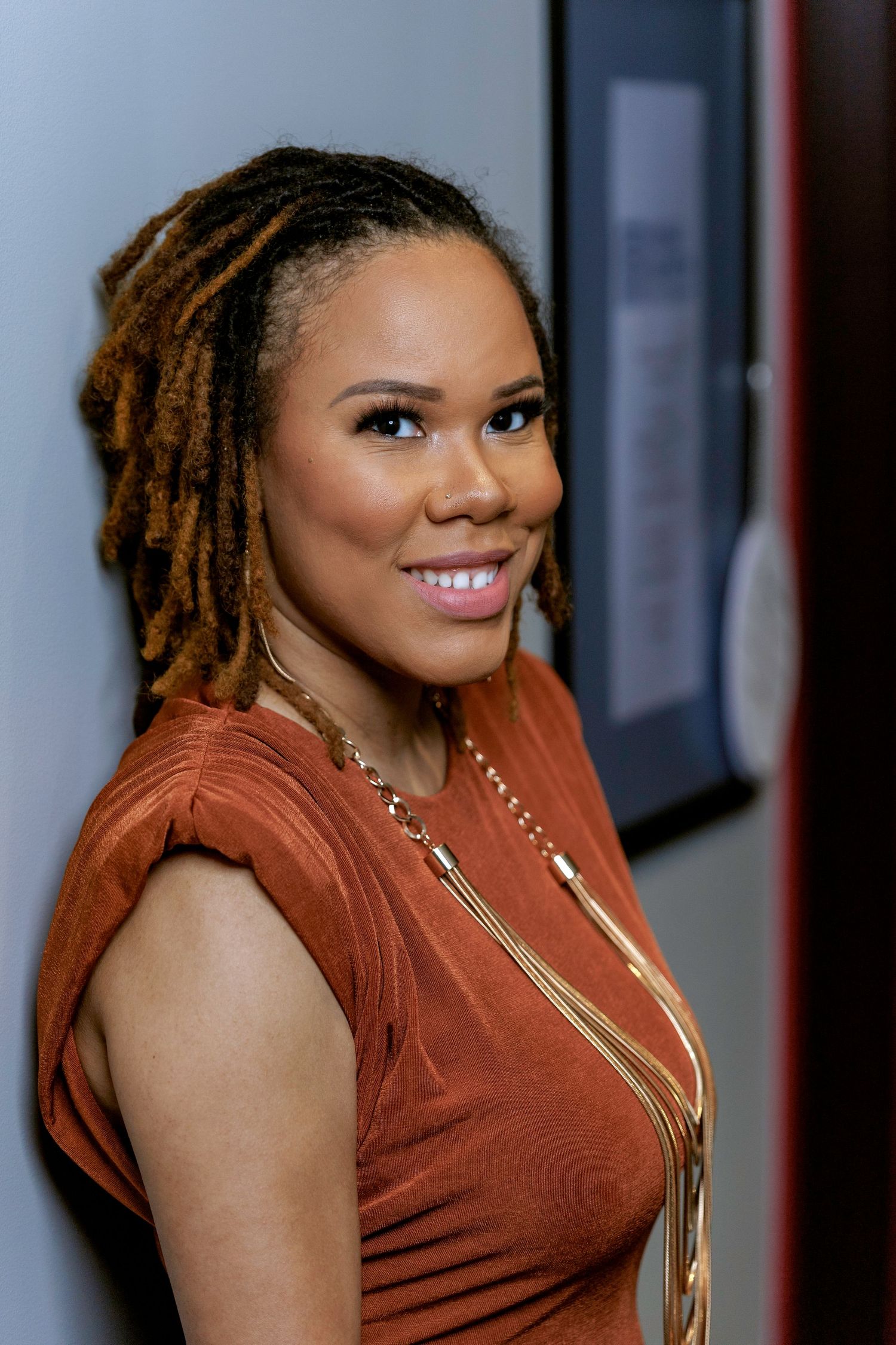 Portrait of a person with dreadlocks wearing a rust-colored top and gold necklace smiling warmly at the camera.