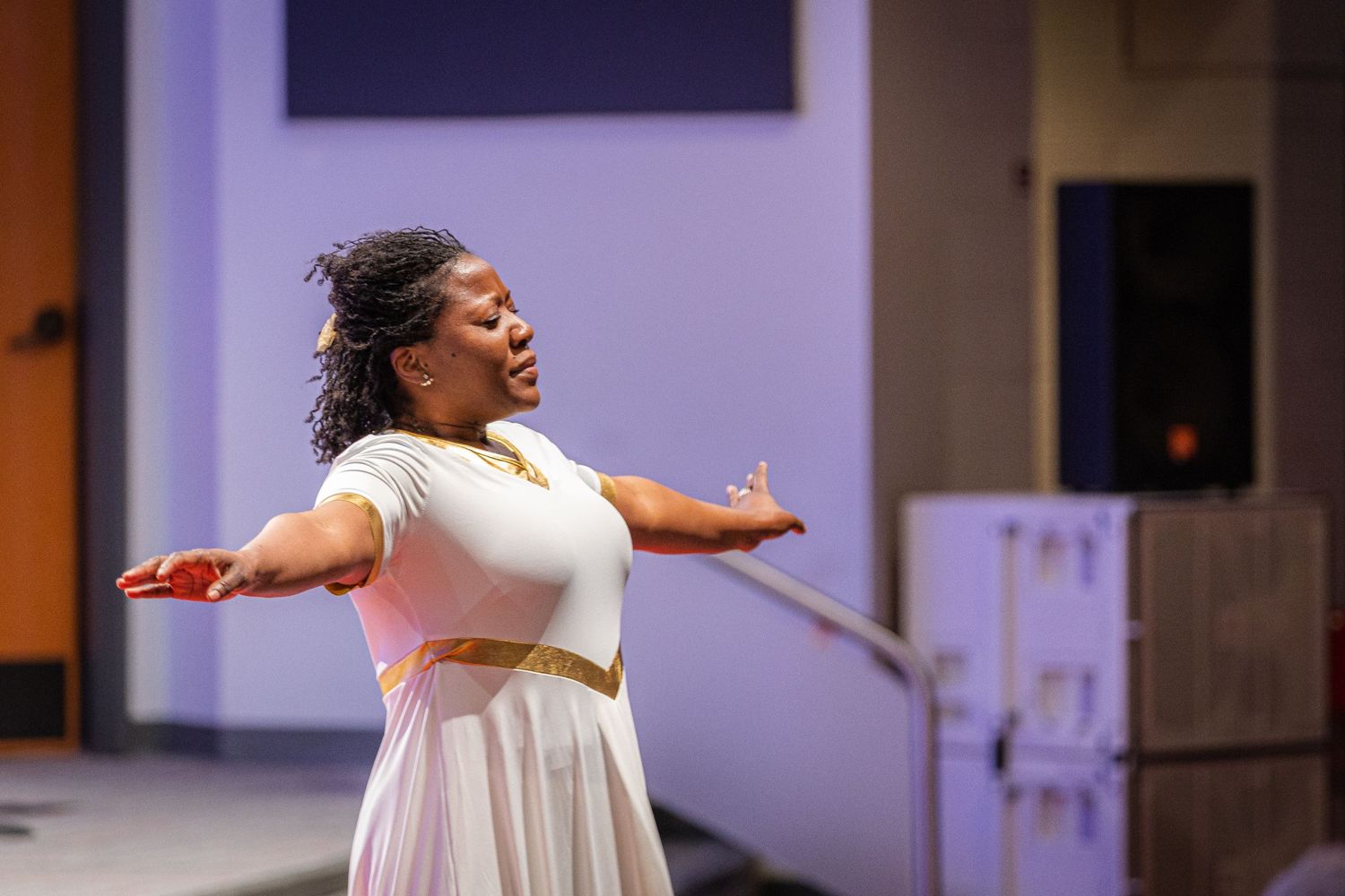 Performer in flowing white dress sings passionately on stage with arms outstretched during theatrical production.