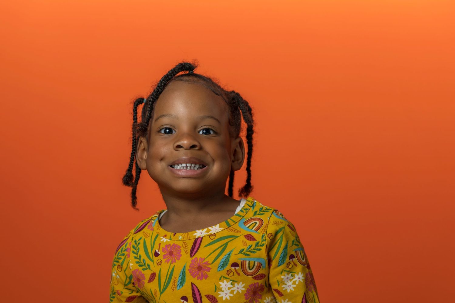 A cheerful child wearing a vibrant yellow floral outfit poses against an orange background.