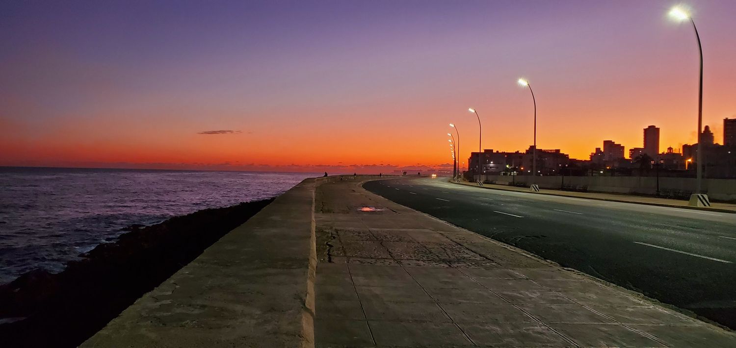 Sunset illuminates a coastal walkway with street lamps and rocks along the shoreline during dusk.