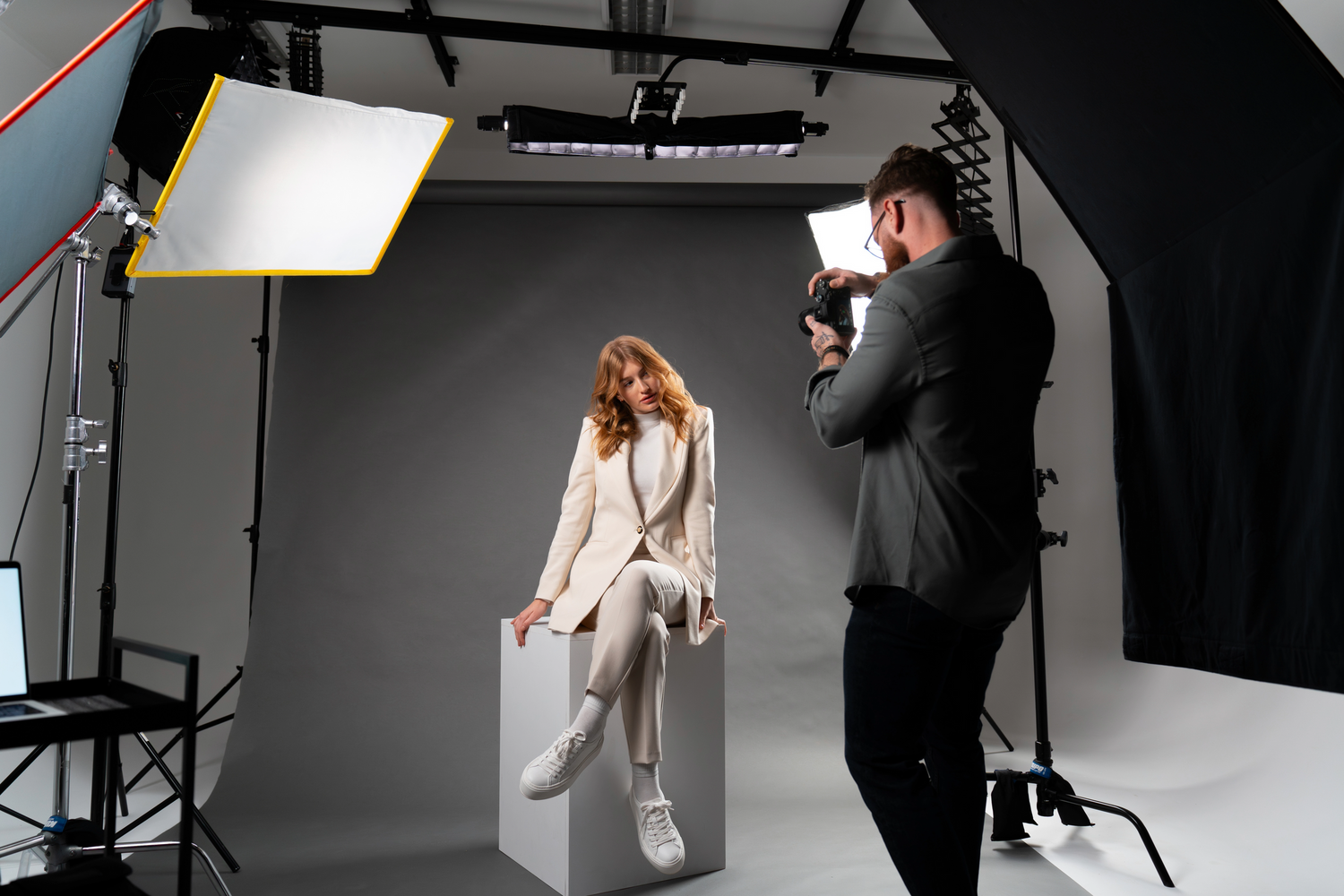 Professional photographer shooting with studio lighting as model poses in white outfit against gray backdrop in photo studio.