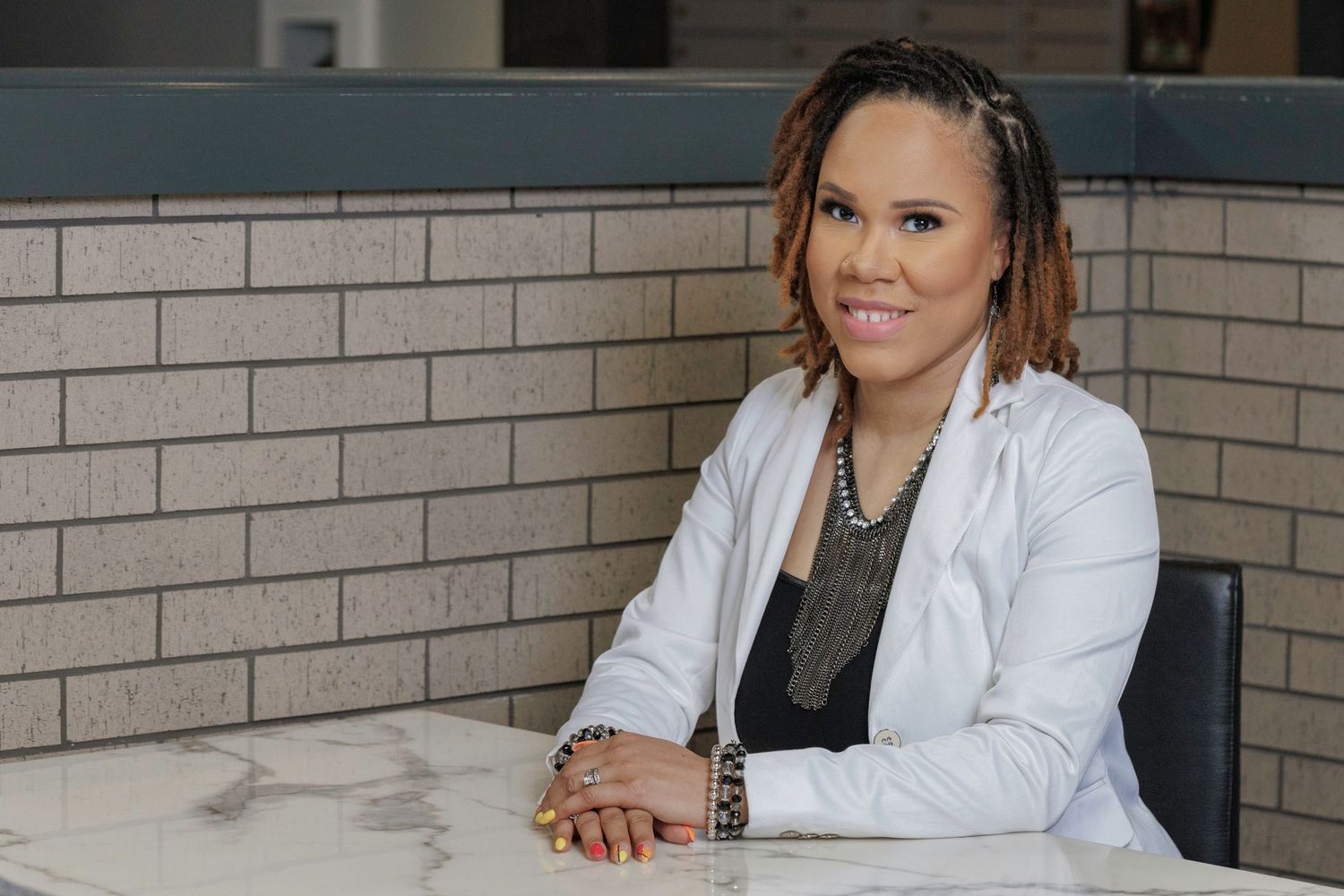 Professional portrait of someone in a white blazer and gray shirt sitting at a desk with a relaxed pose and friendly expression.