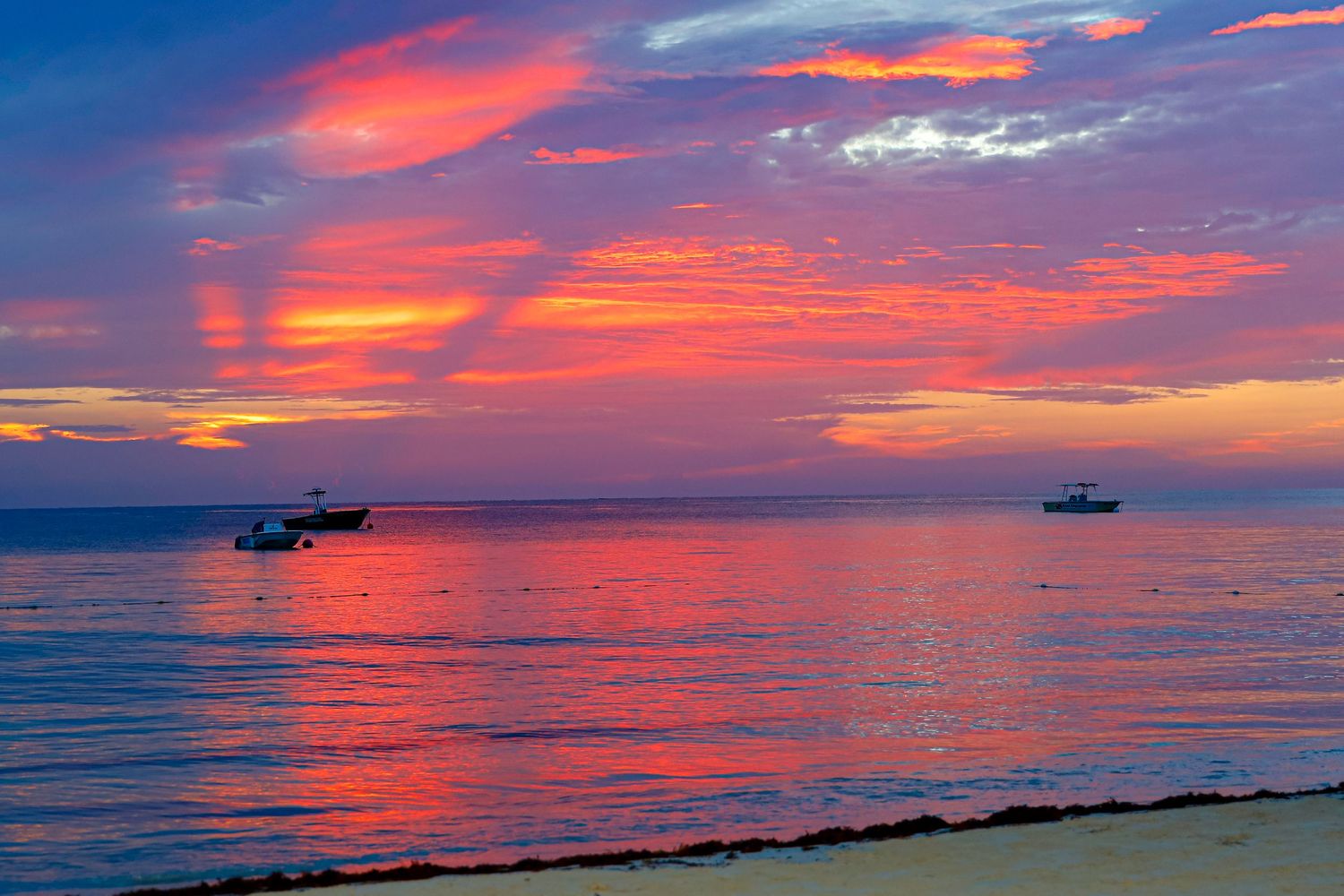 Dramatic orange and pink sunset sky reflects on calm ocean waters with silhouettes of boats on the horizon.