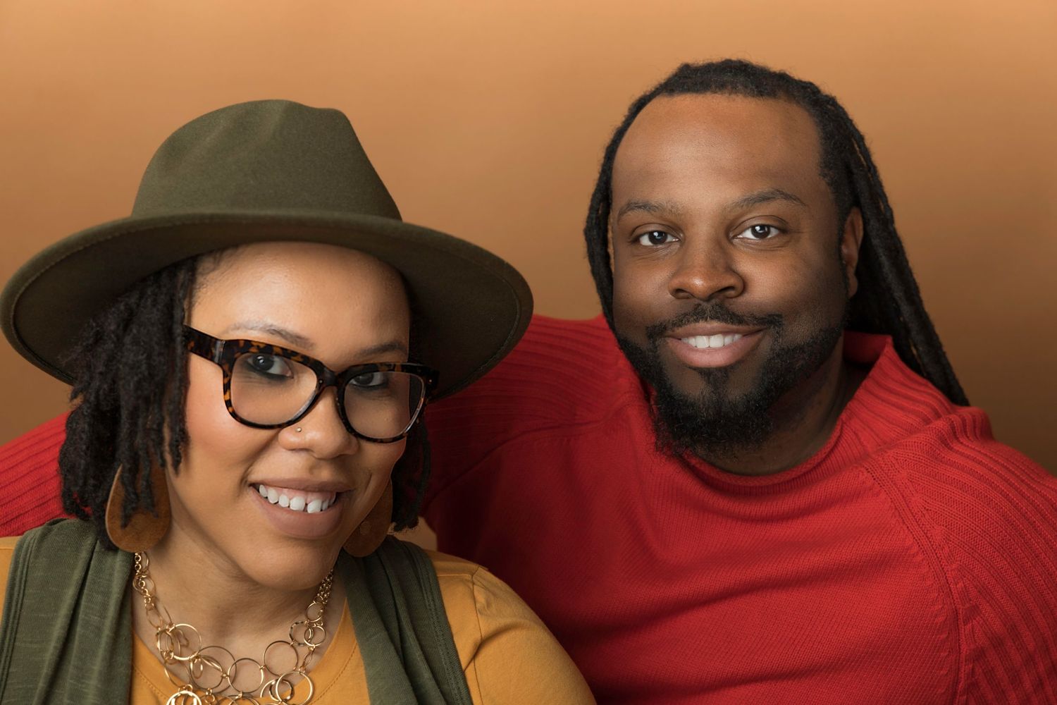Stylish couple smiling in casual portrait against warm brown background.