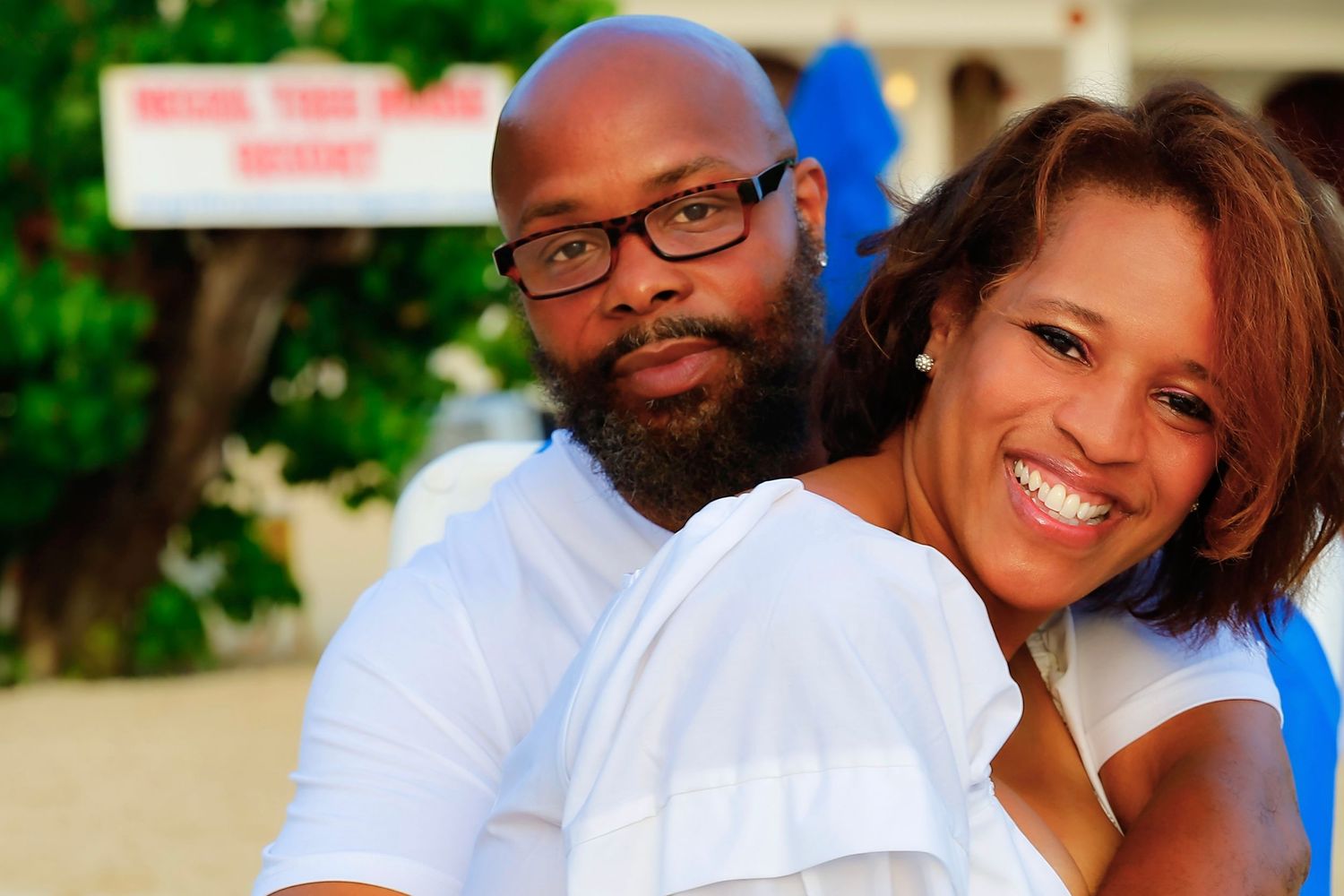 A happy couple in casual white attire sits close together and smiles warmly at the camera outdoors.