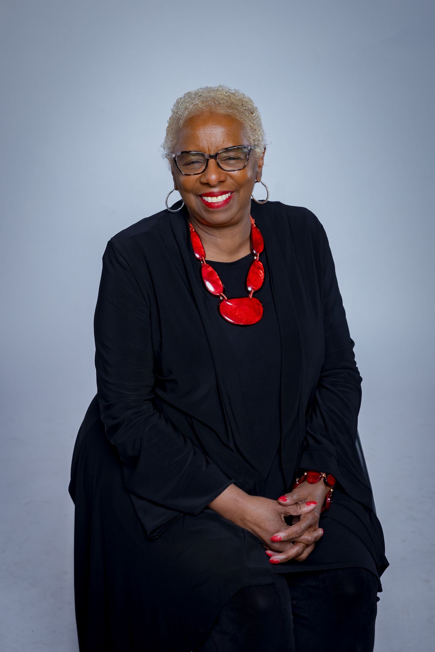 A woman in black attire with a red necklace poses elegantly in a studio setting.