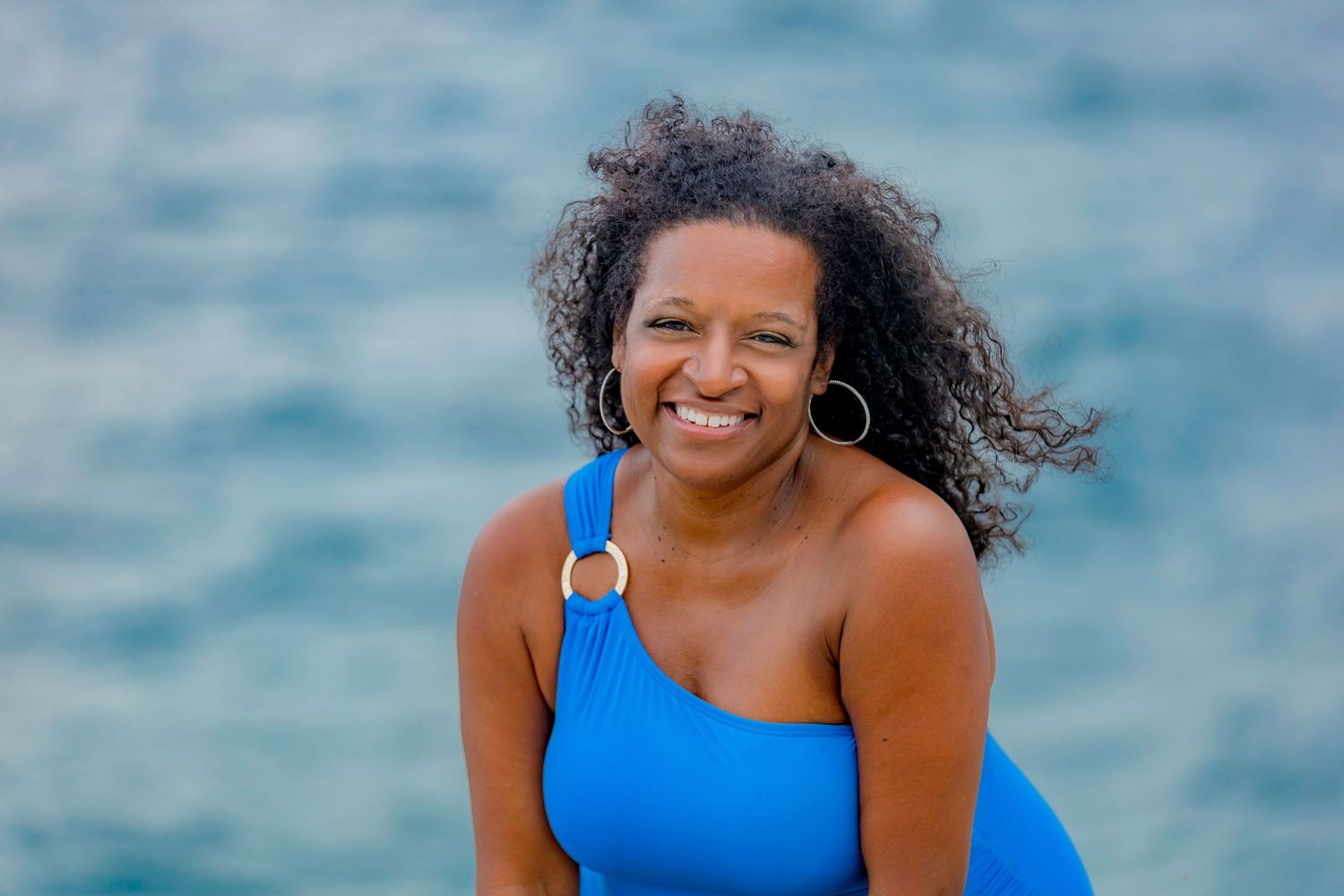 Woman in blue swimsuit smiles joyfully by ocean.
