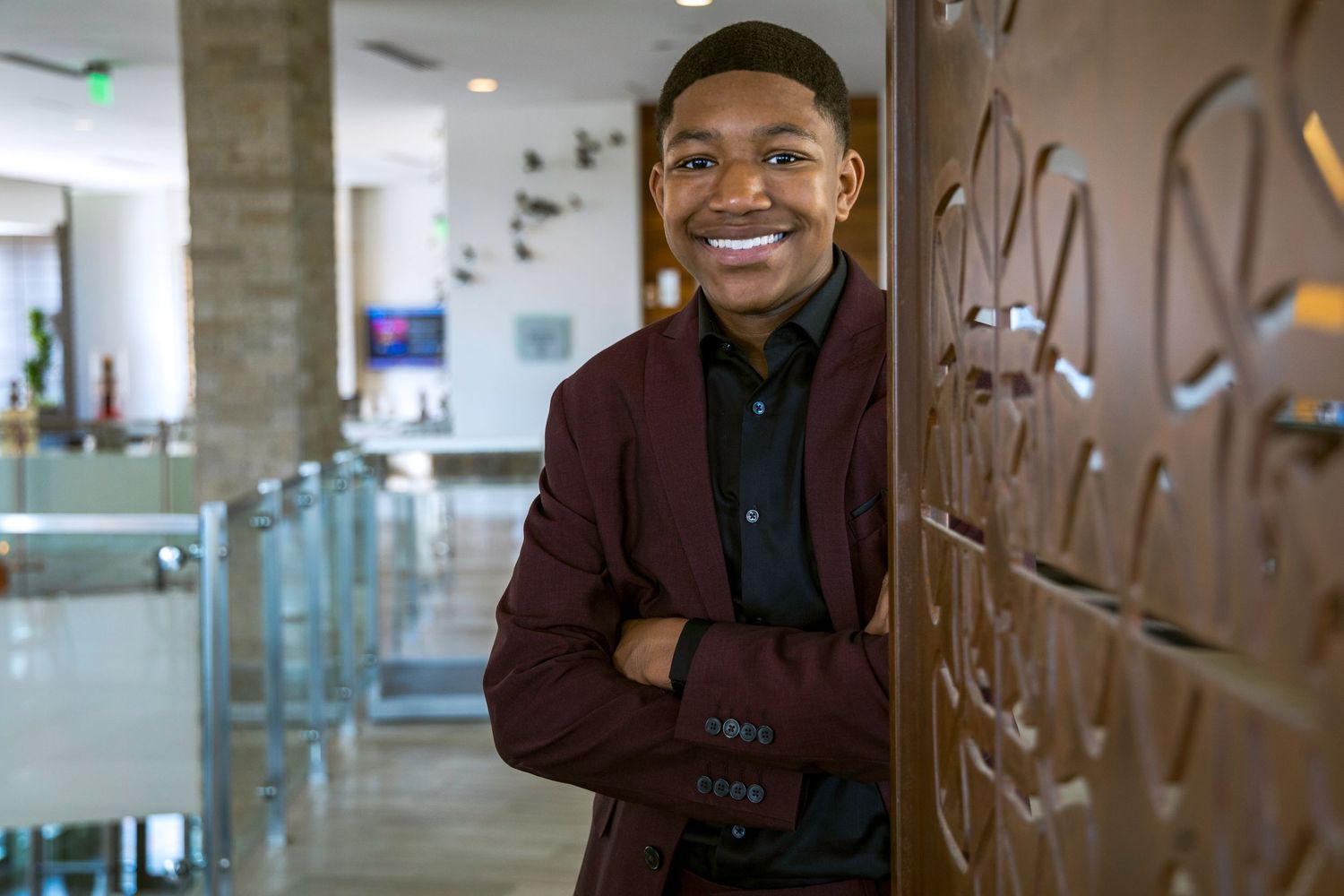 Young man in burgundy blazer leans against decorative wall in modern office space.