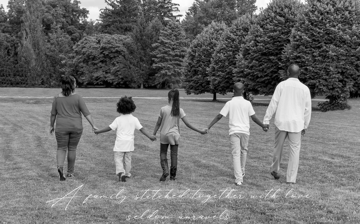 A family holding hands while walking away from the camera across a grassy park in black and white.